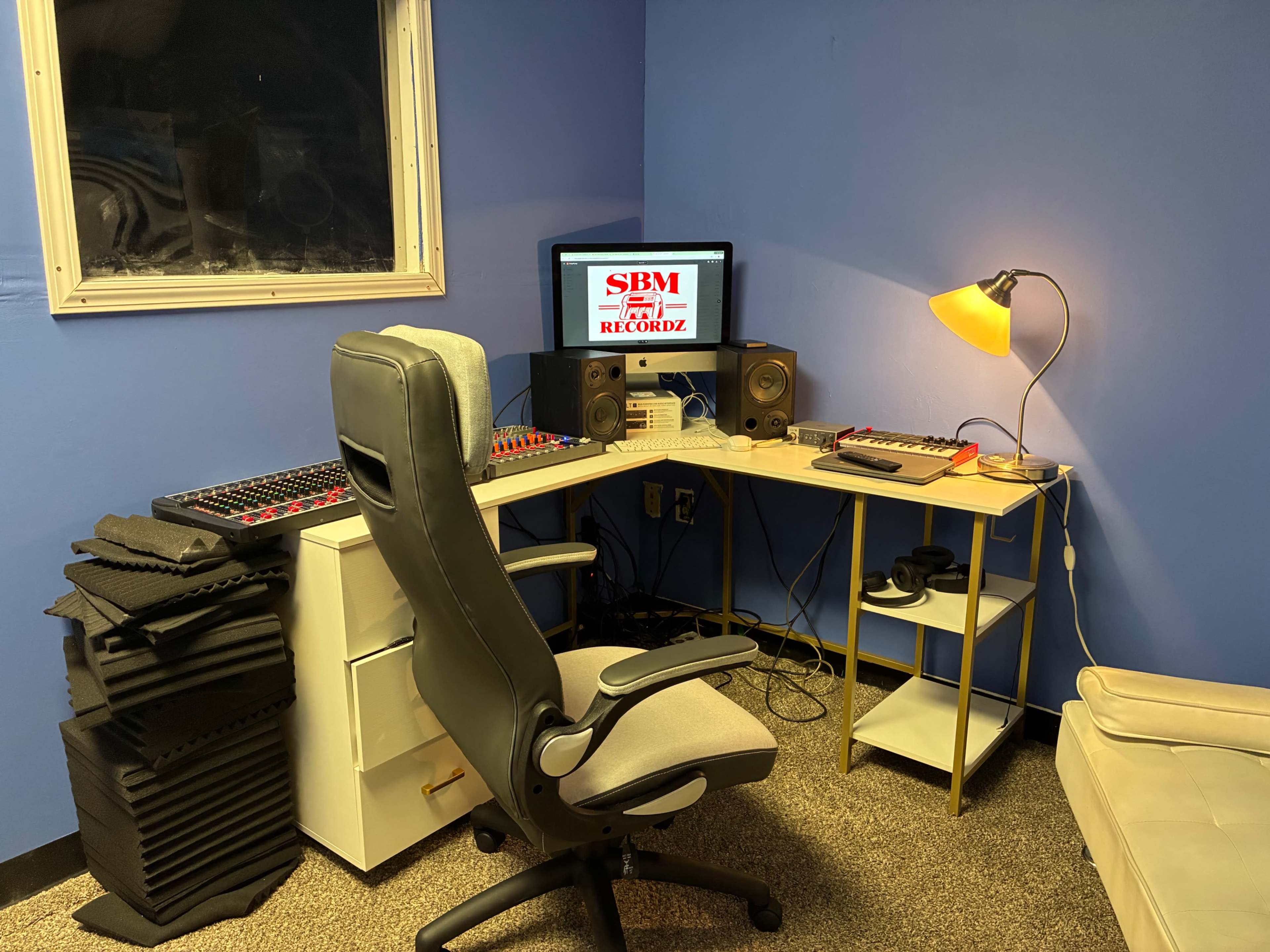 The image shows a compact recording studio featuring a computer desk with a monitor, speakers, a MIDI controller, and an adjustable chair, all set against a blue wall.