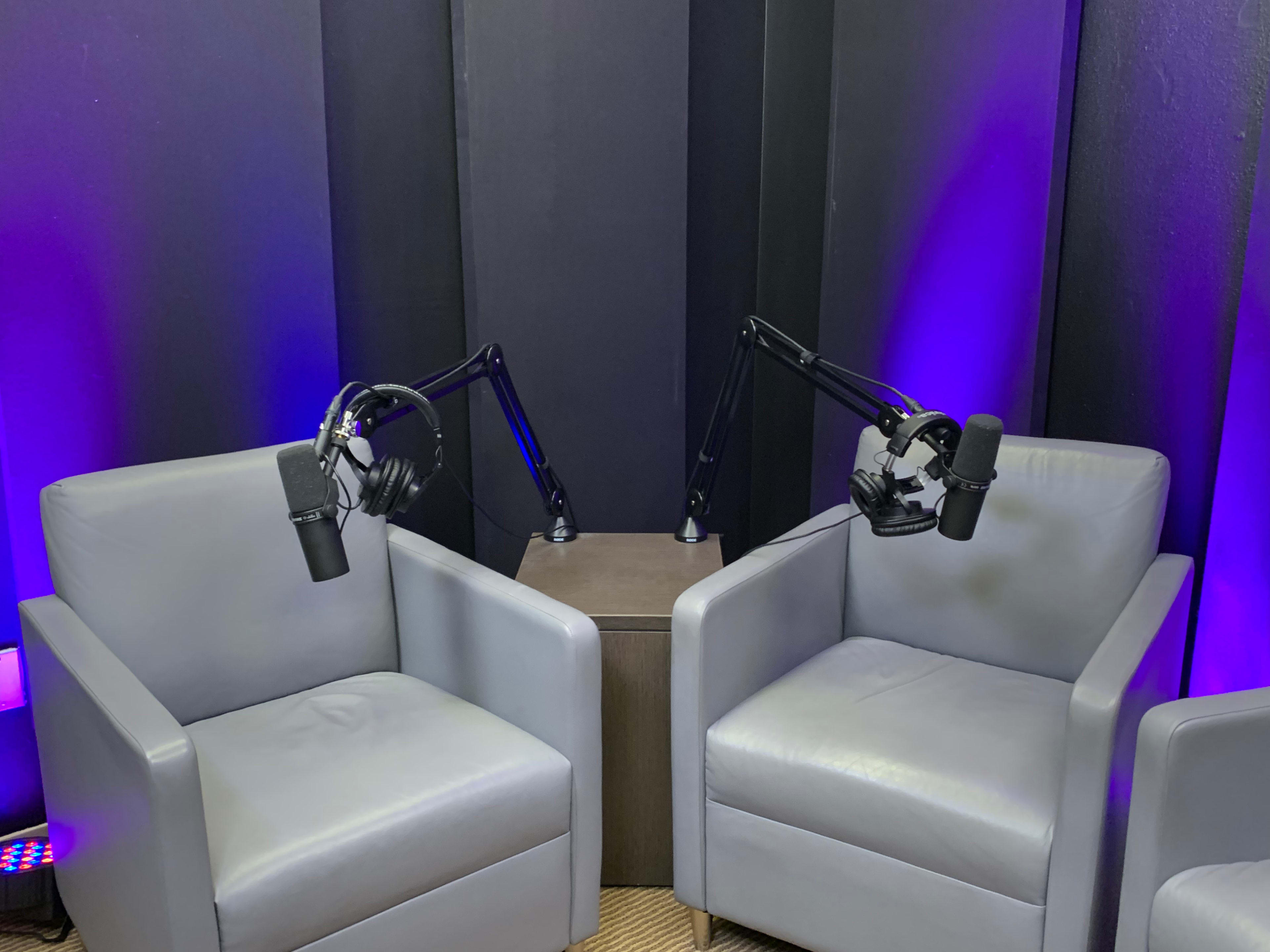 Two gray armchairs are positioned facing each other under a backdrop of purple lighting, each equipped with a microphone on an adjustable arm.