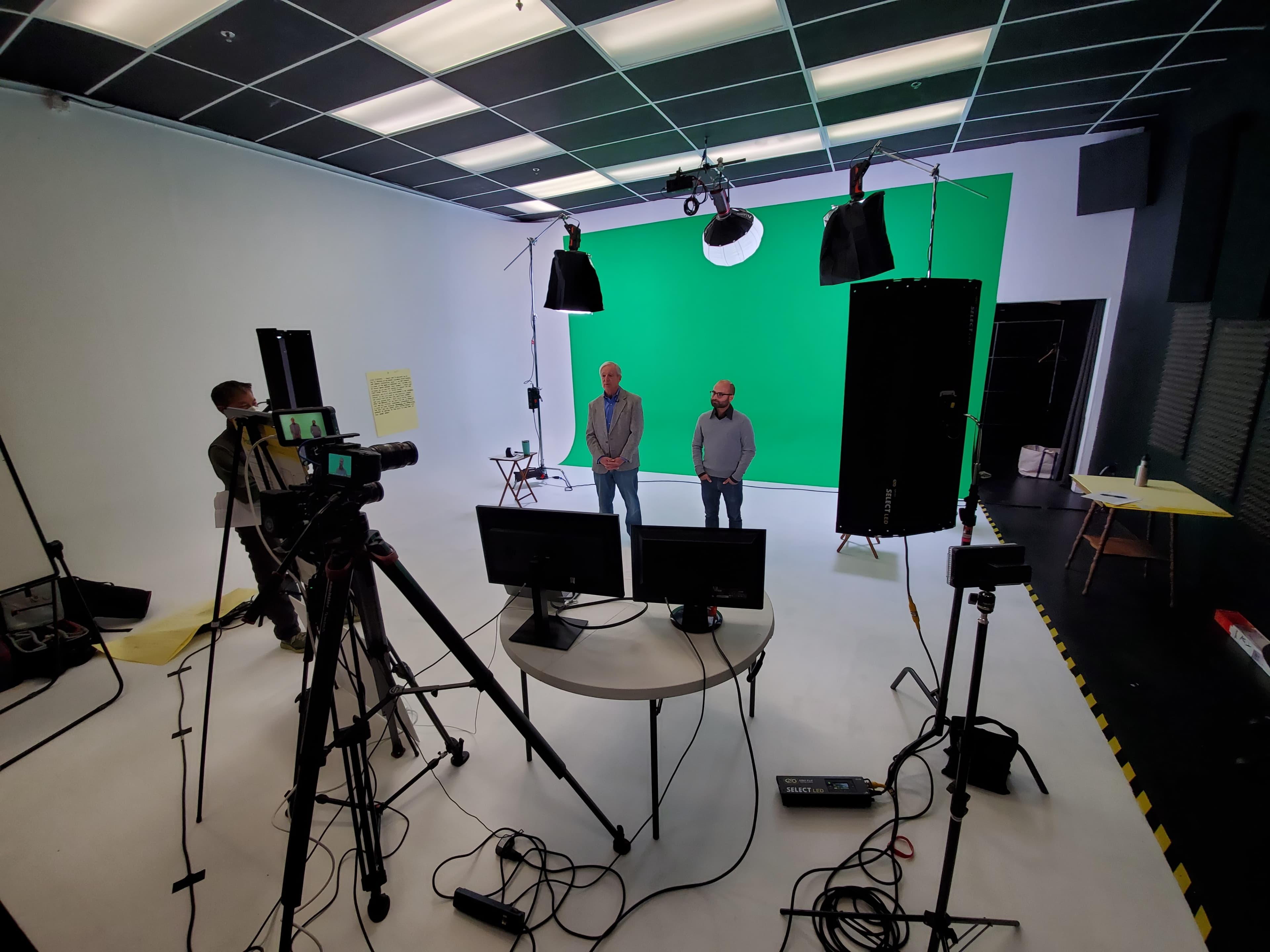 Two individuals stand on a stage with a green screen backdrop, surrounded by cameras and studio lighting equipment.