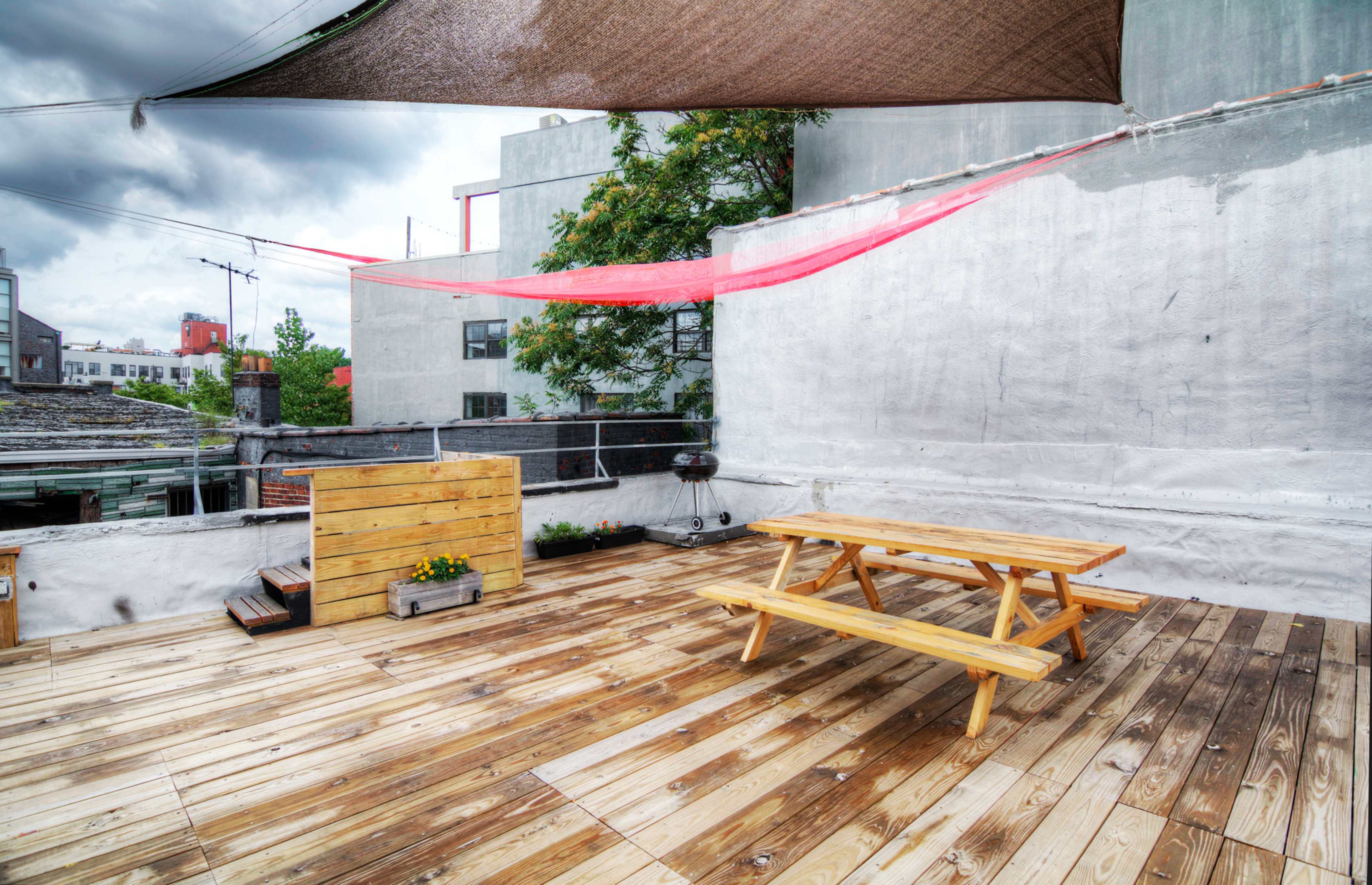 A rooftop deck with wooden flooring, a picnic table, and shaded areas created by large fabric drapes, surrounded by buildings and greenery.
