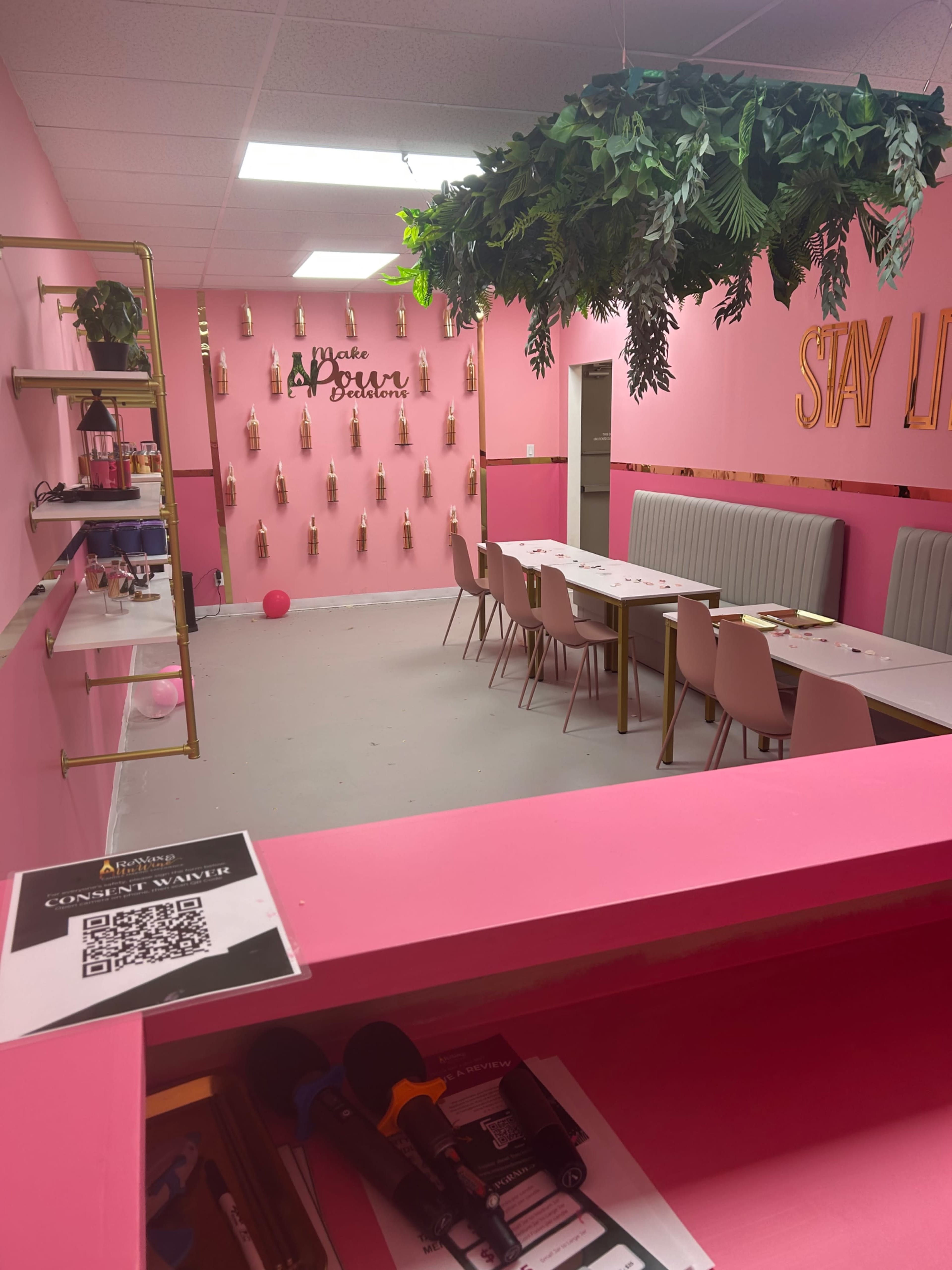 The image shows a vibrant, pink-themed interior space with a seating area featuring tables and chairs, decorative plants hanging from the ceiling, and a wall adorned with bottles and the words "Stay Loved."