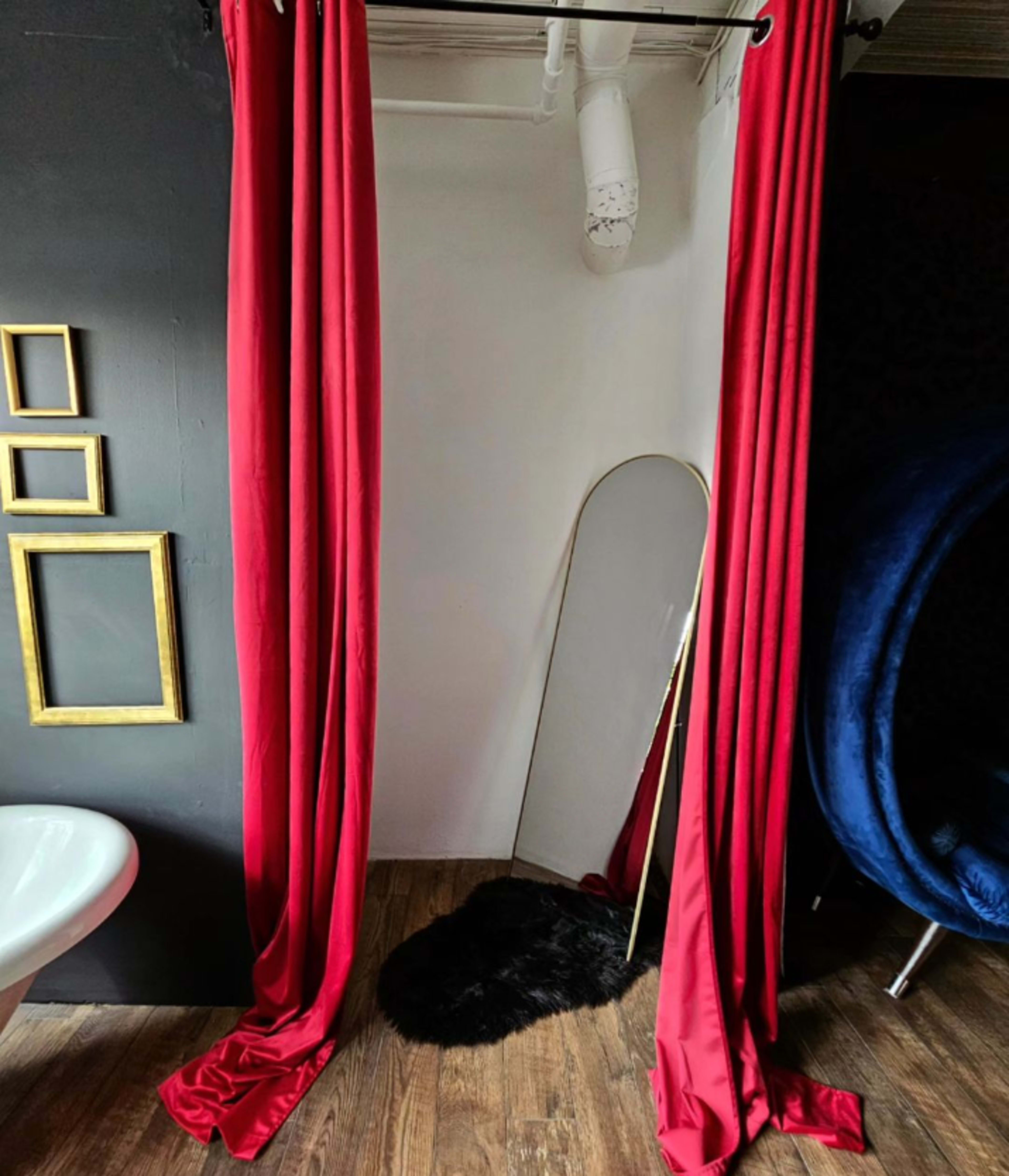 The image shows a corner of a room with red curtains draping from a rod, a large mirror leaning against the wall, and a dark fur rug on the floor.