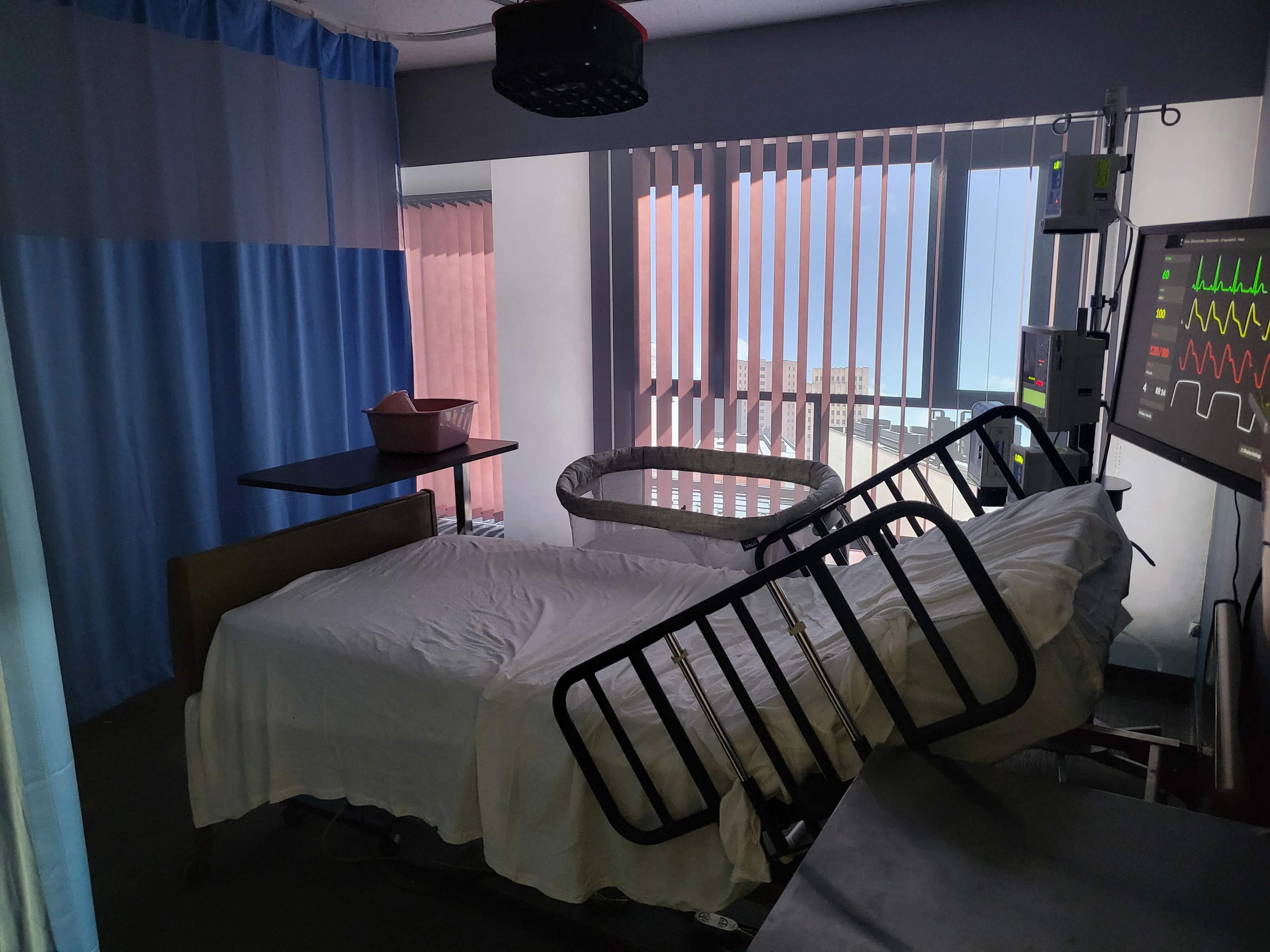 The image shows an empty hospital room with a partially made bed, a monitor displaying medical signals, and curtains separating the space.