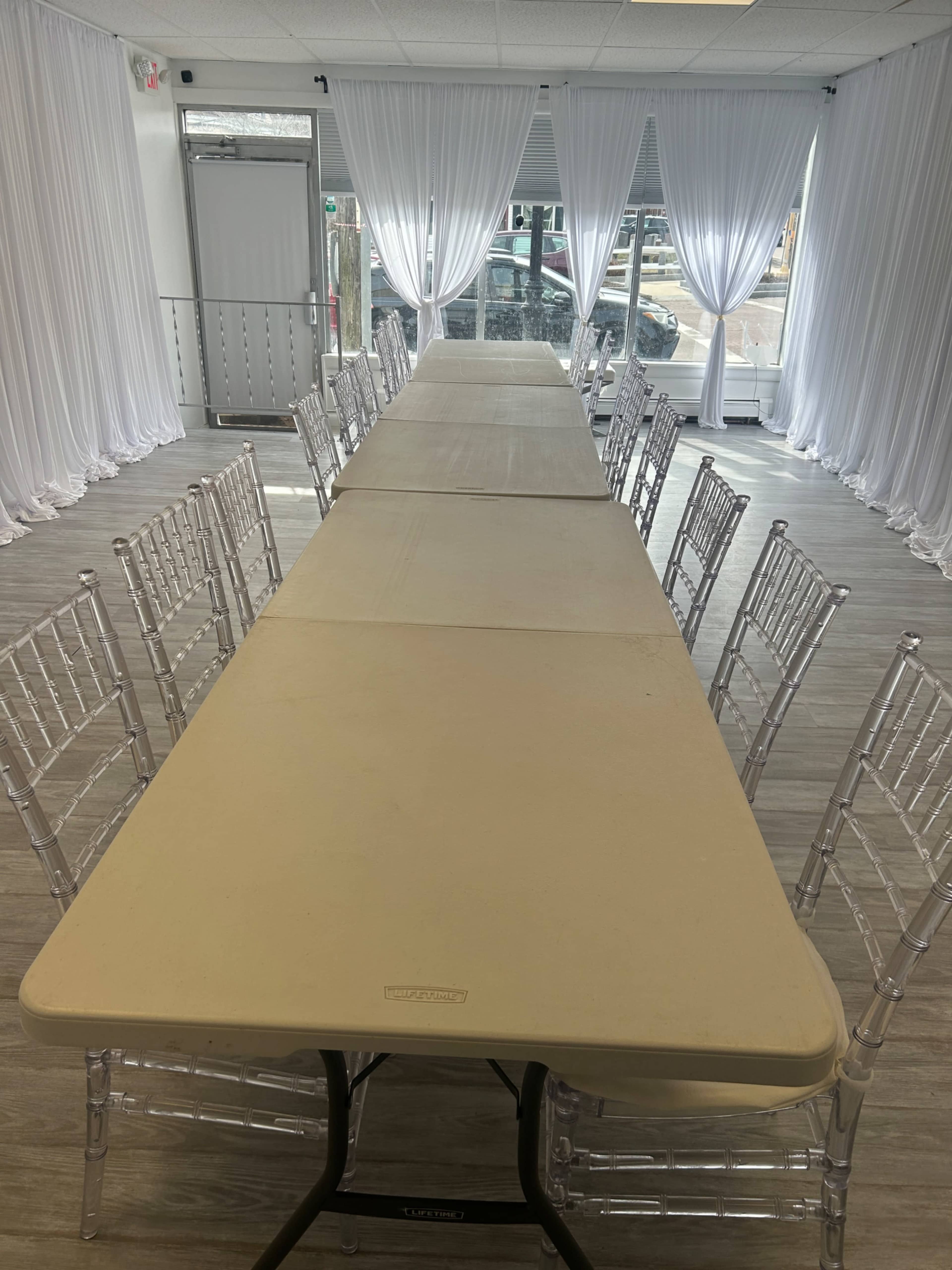 A long table is set up in a well-lit room with white drapes and transparent chairs.