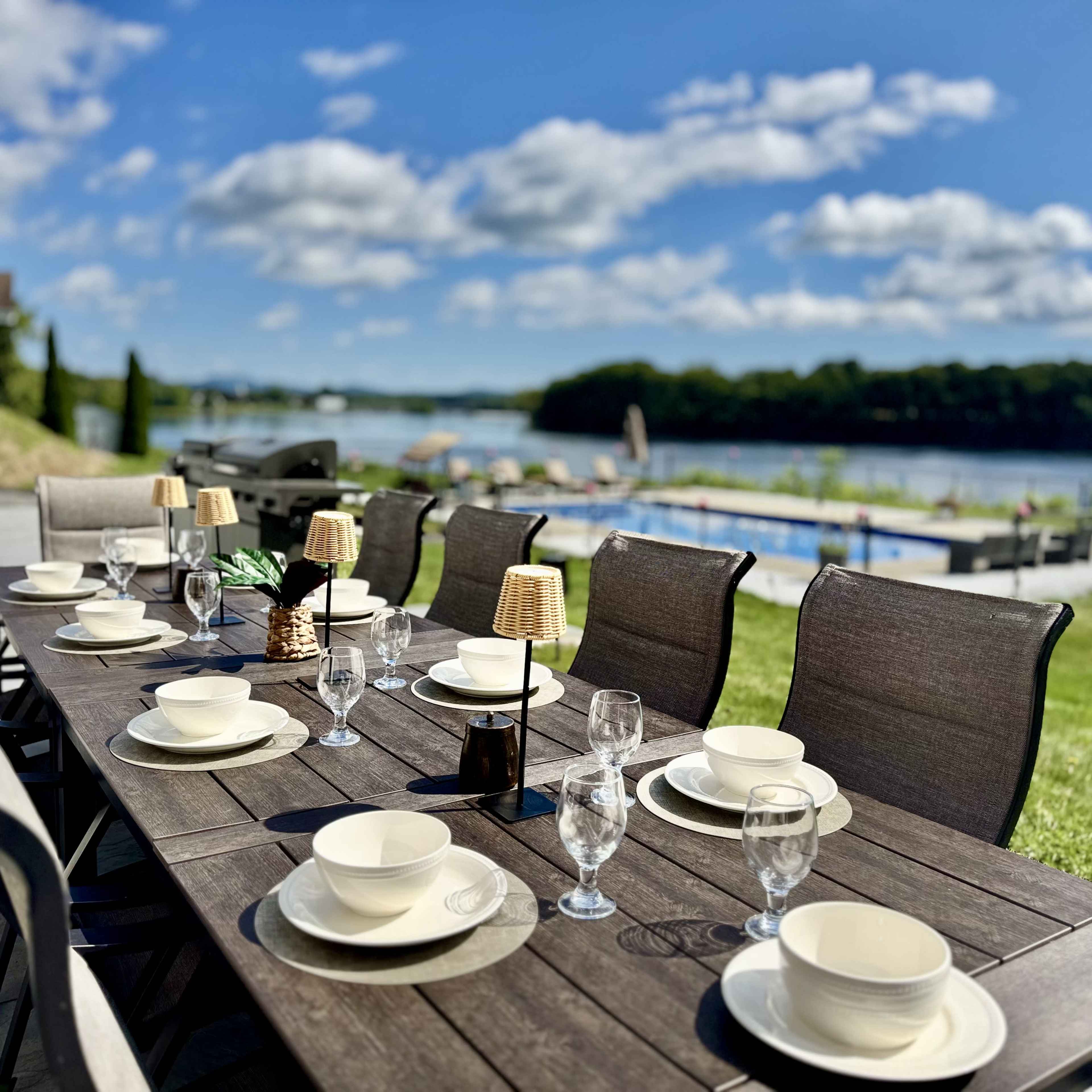A long outdoor dining table is set with plates and glasses, overlooking a swimming pool and a body of water under a blue sky with clouds.
