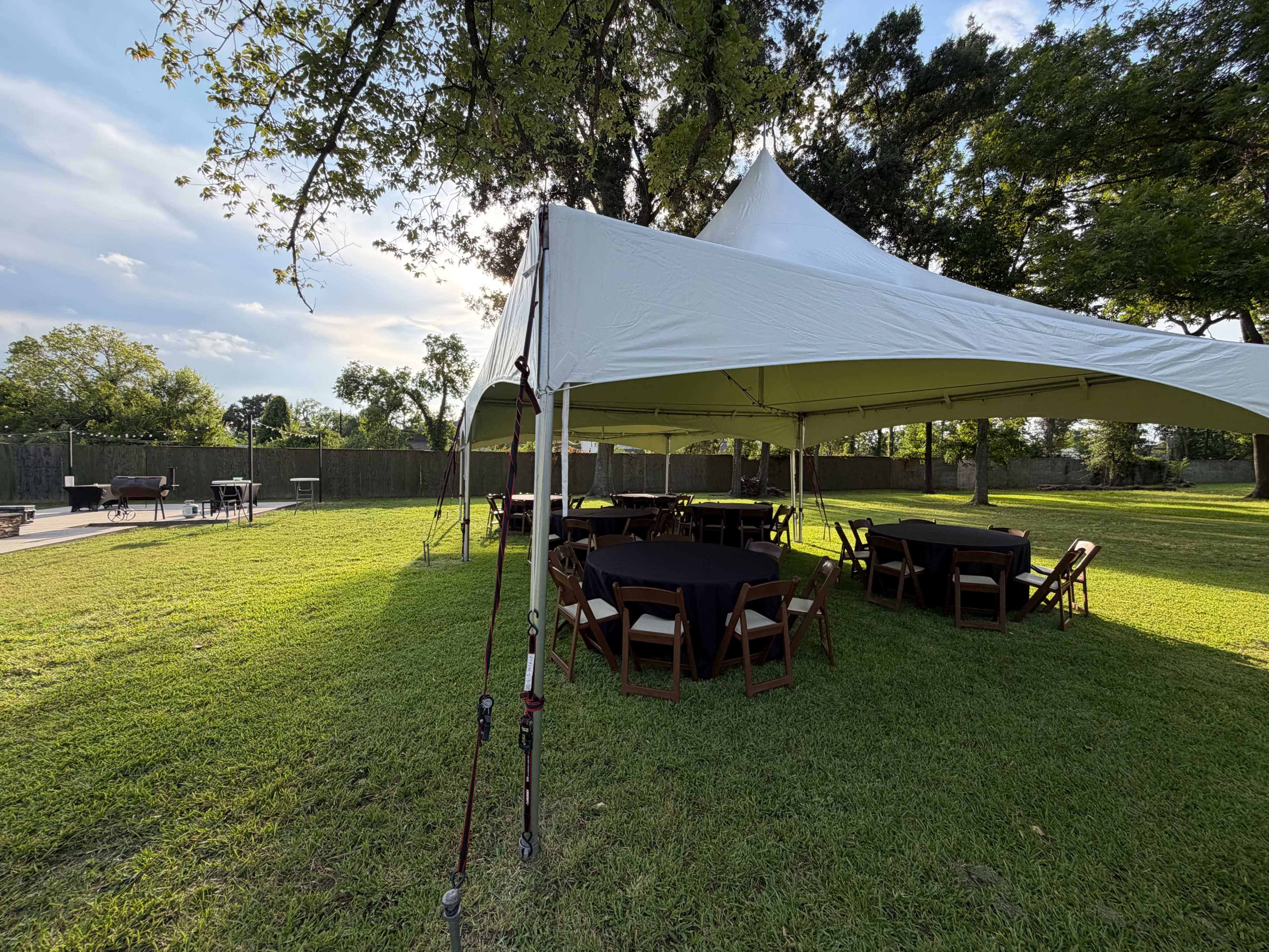 A large white tent with round tables and chairs is set up on a grassy outdoor area, surrounded by trees and a fence, with a patio and grilling area in the background.