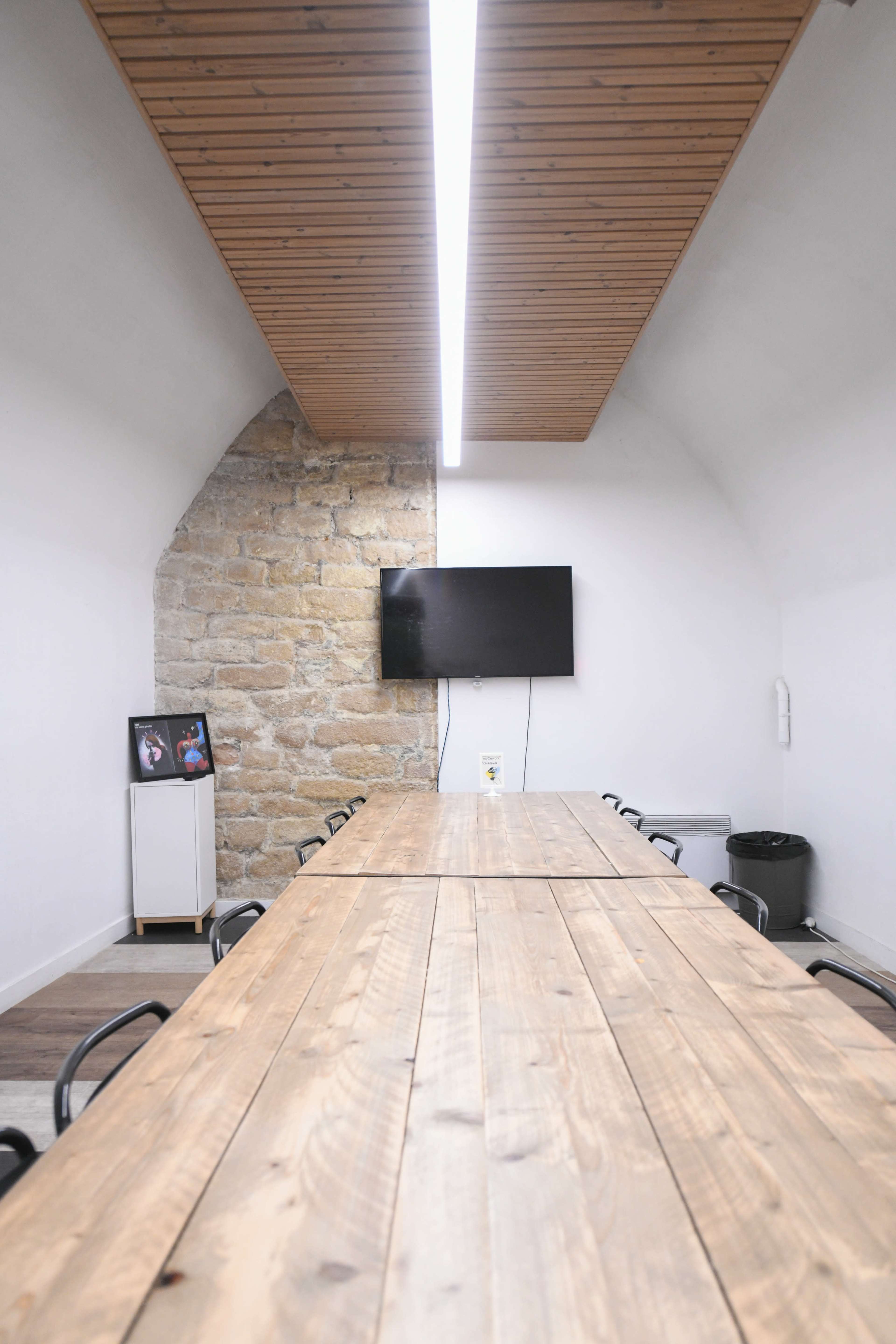 A long wooden table is set in a conference room with a stone wall on one side and a TV mounted on the opposite wall.