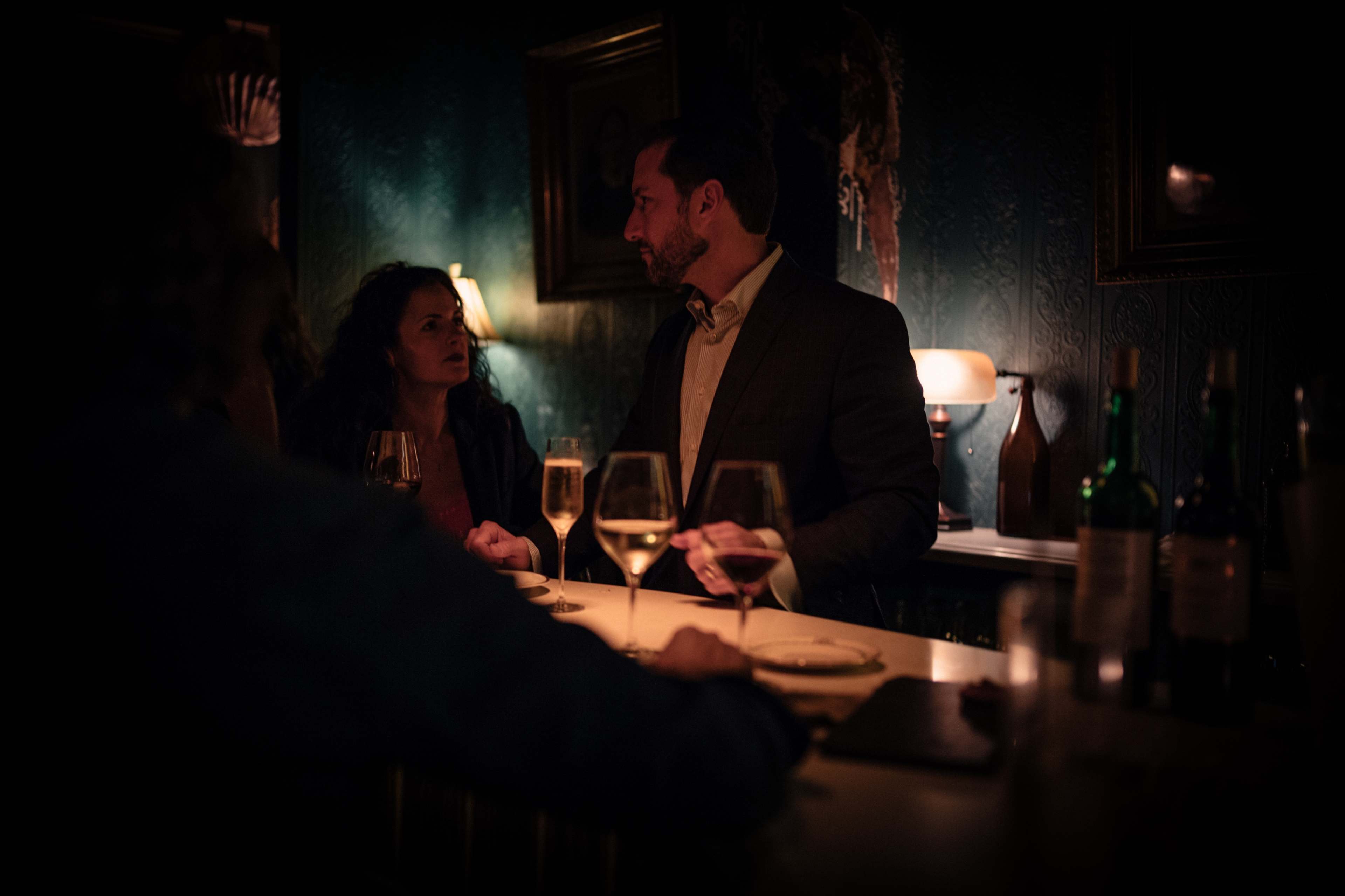 A man and a woman are engaged in conversation at a dimly lit bar, with glasses of champagne and wine on the countertop.