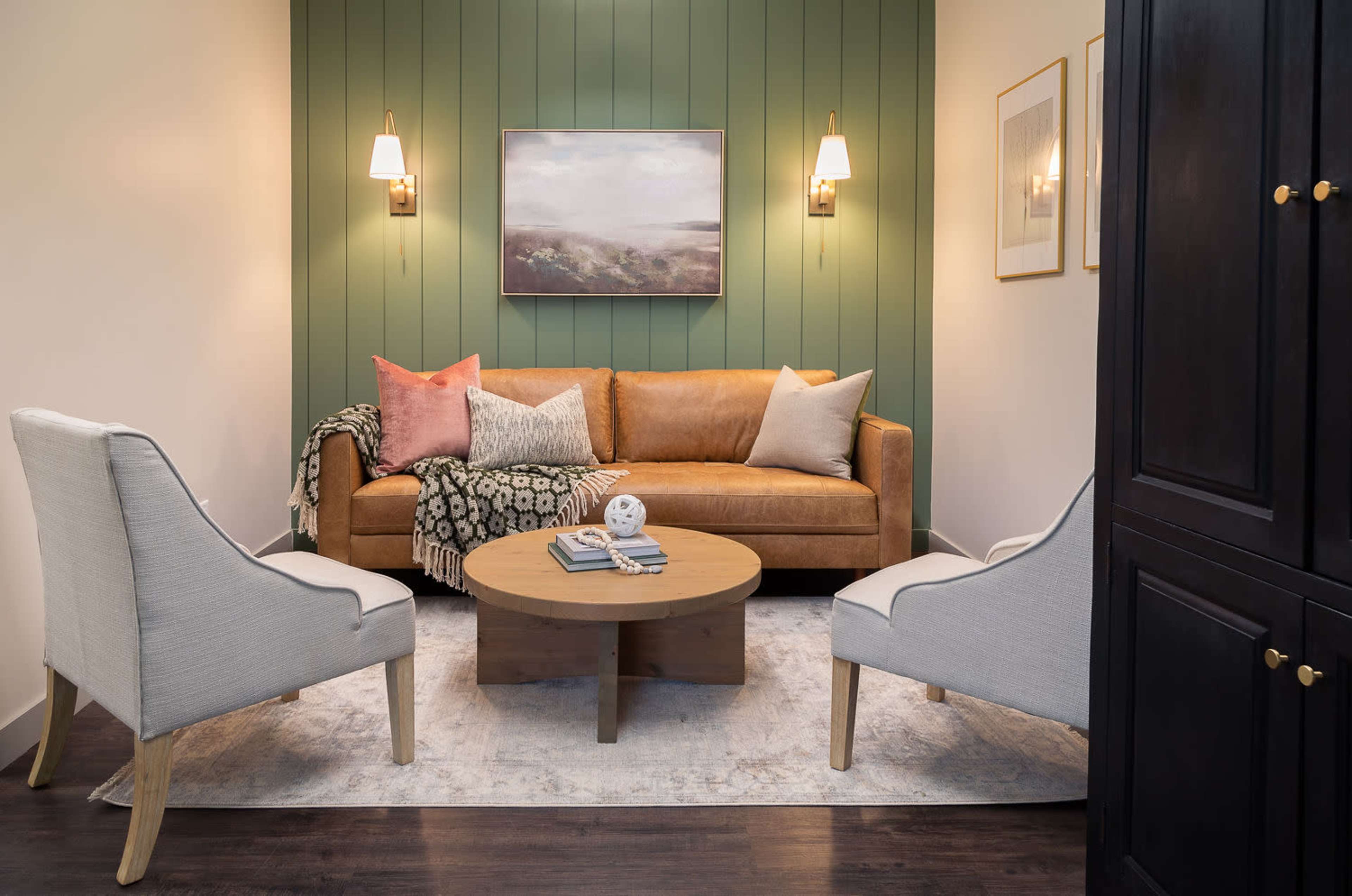 A cozy living room features a brown leather sofa with decorative pillows, flanked by two light-colored chairs and a round wooden coffee table on a patterned rug.