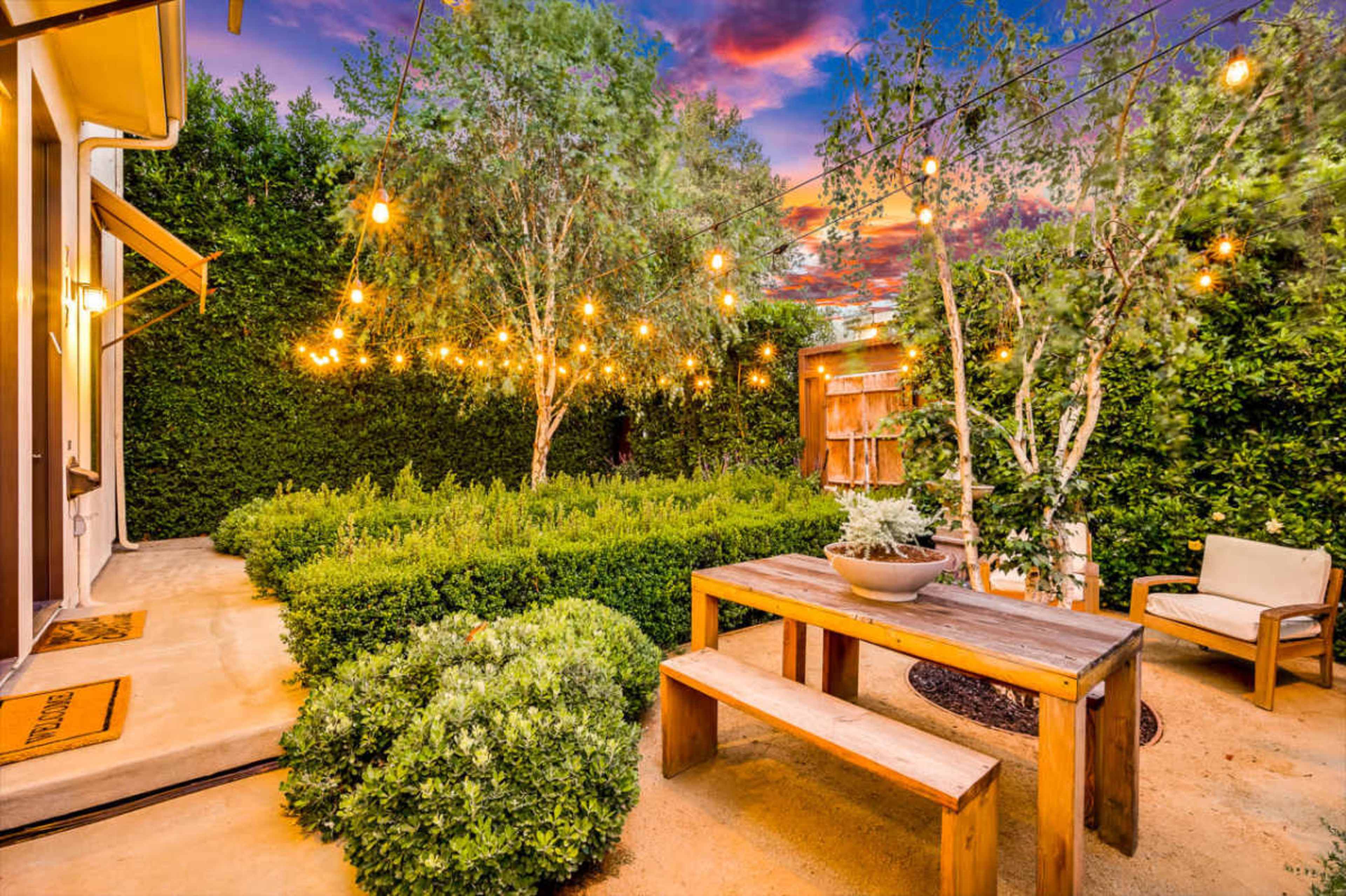The image shows a landscaped garden with string lights overhead, featuring a wooden table and seating area surrounded by green shrubbery and trees.