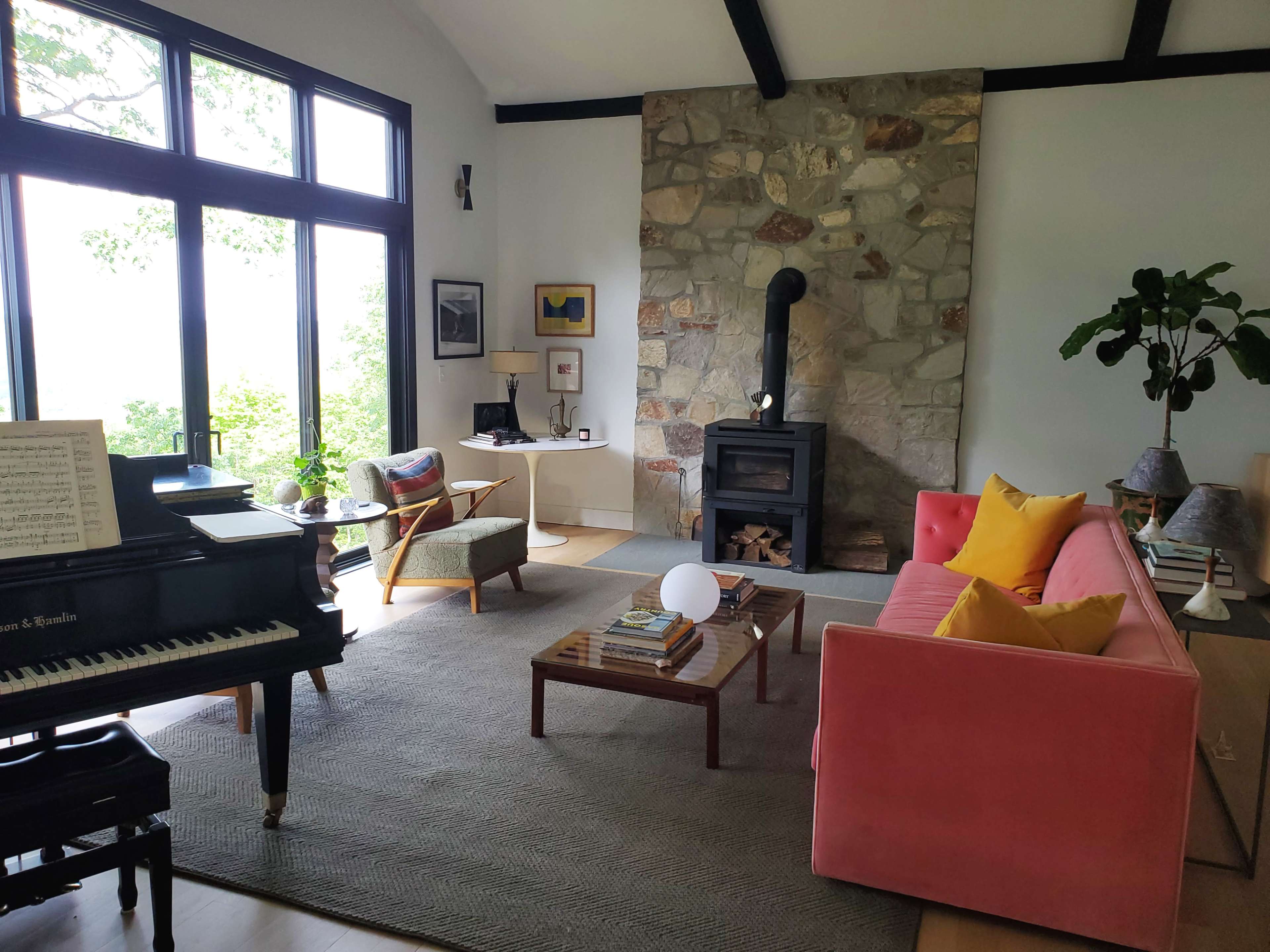 The image features a cozy living room with a pink sofa, a piano, a stone fireplace, and large windows allowing natural light.