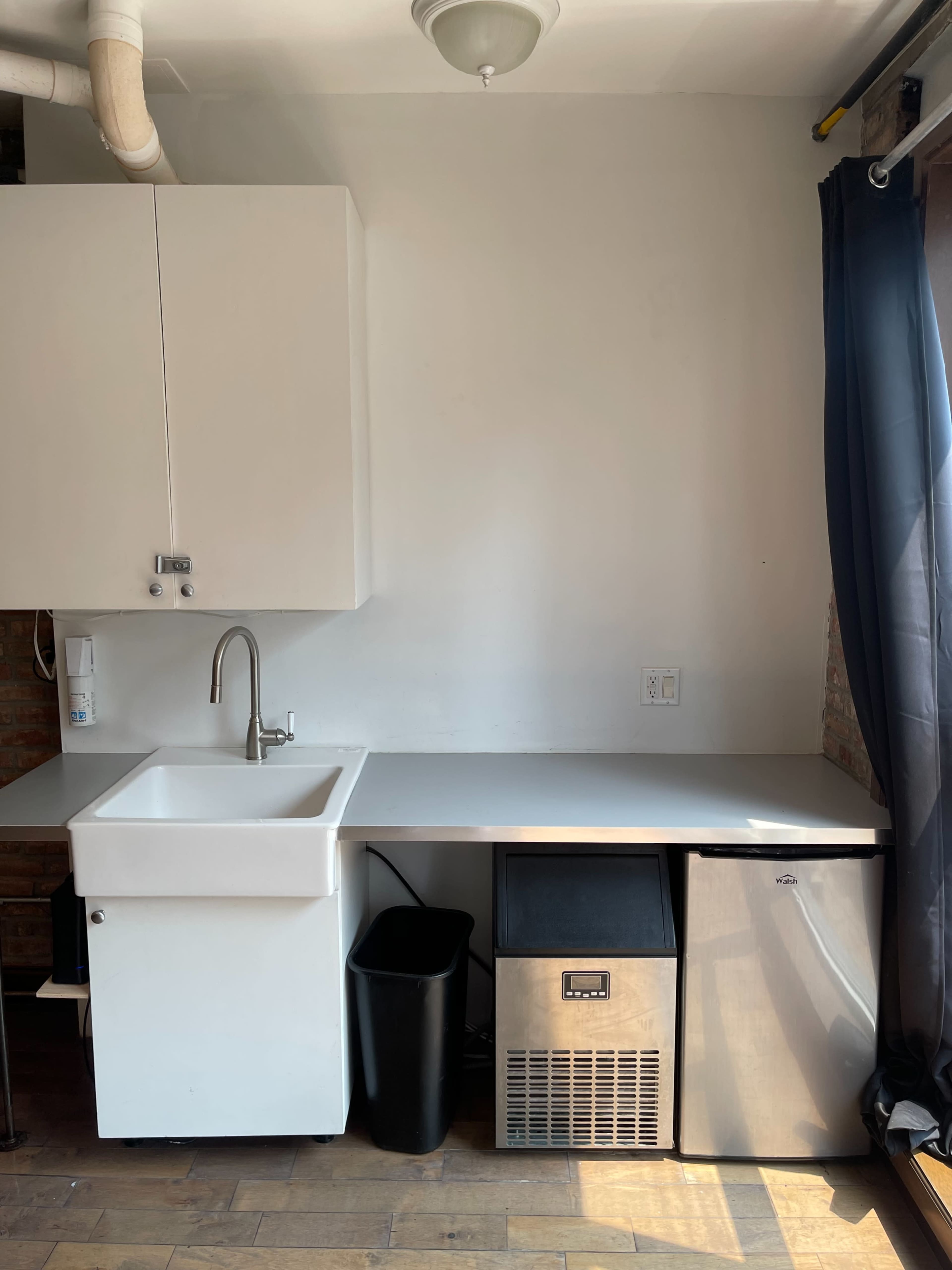 A minimalist kitchen area featuring a sink, a small ice maker, a metal refrigerator, and a black trash can against a light-colored wall.