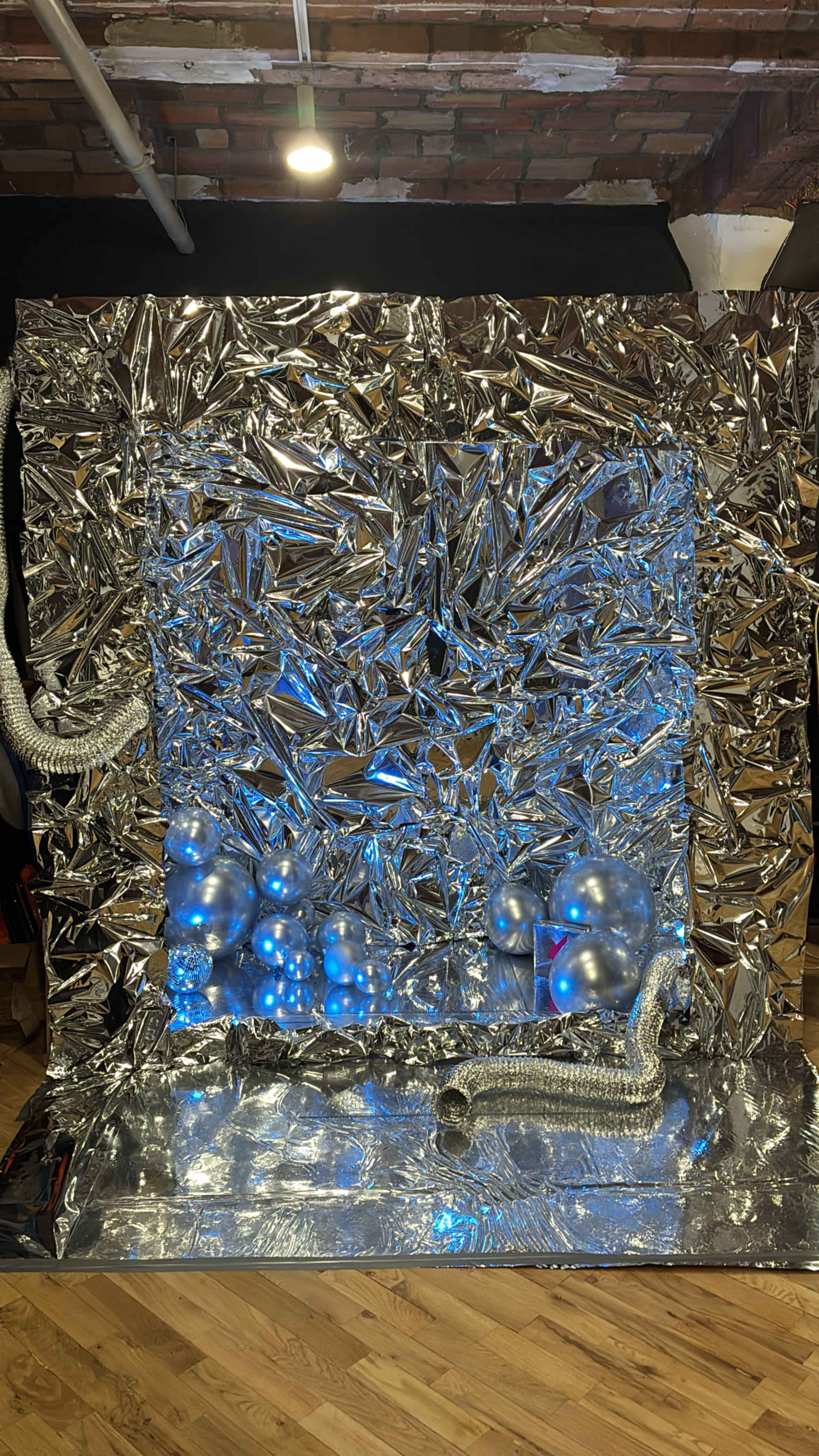 The image shows a large, reflective installation made of crumpled silver material, featuring blue lights and several inflated balloons arranged at the bottom.