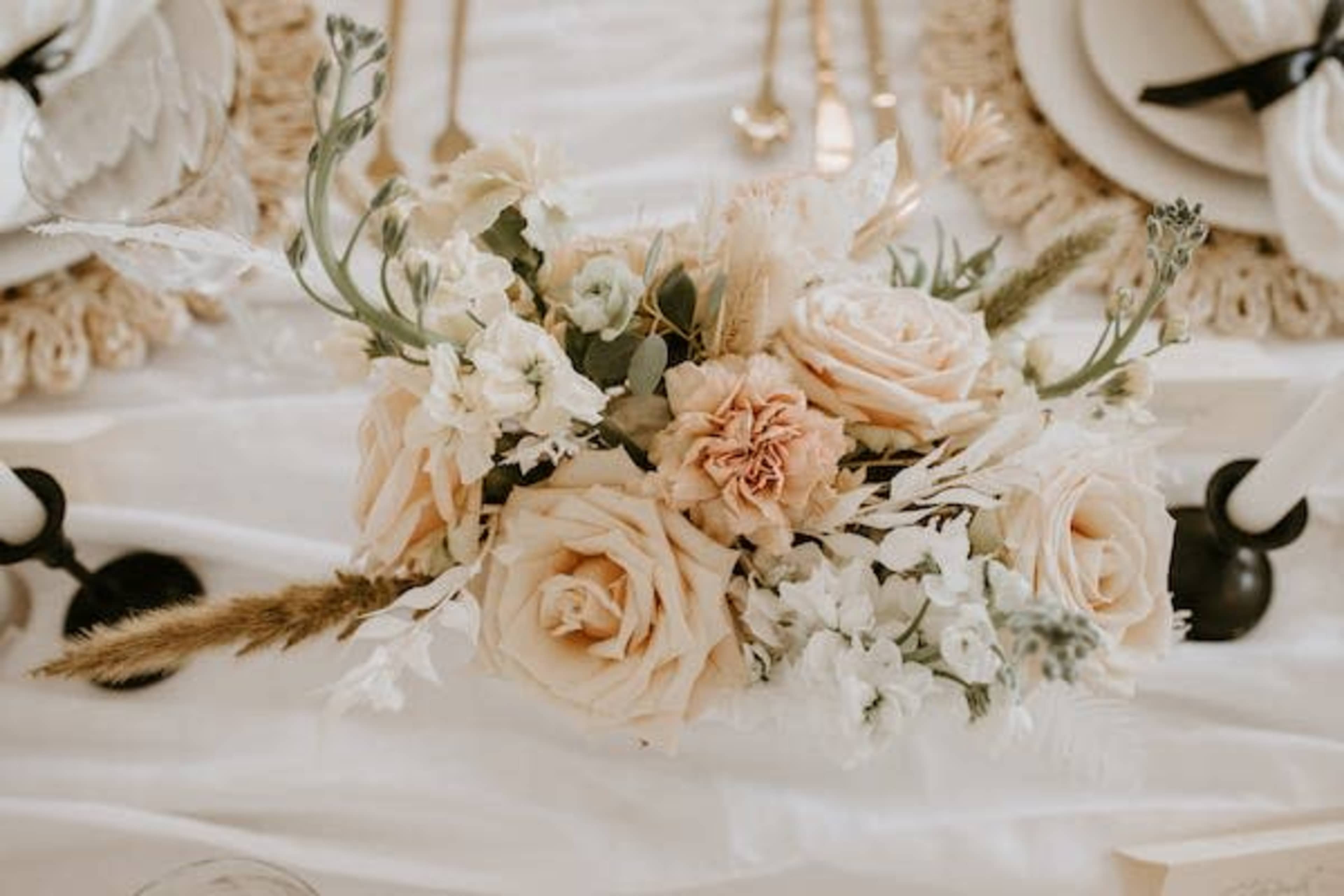 A floral centerpiece with beige and white roses, along with assorted greenery, is placed on a table set with elegant gold utensils and plates.