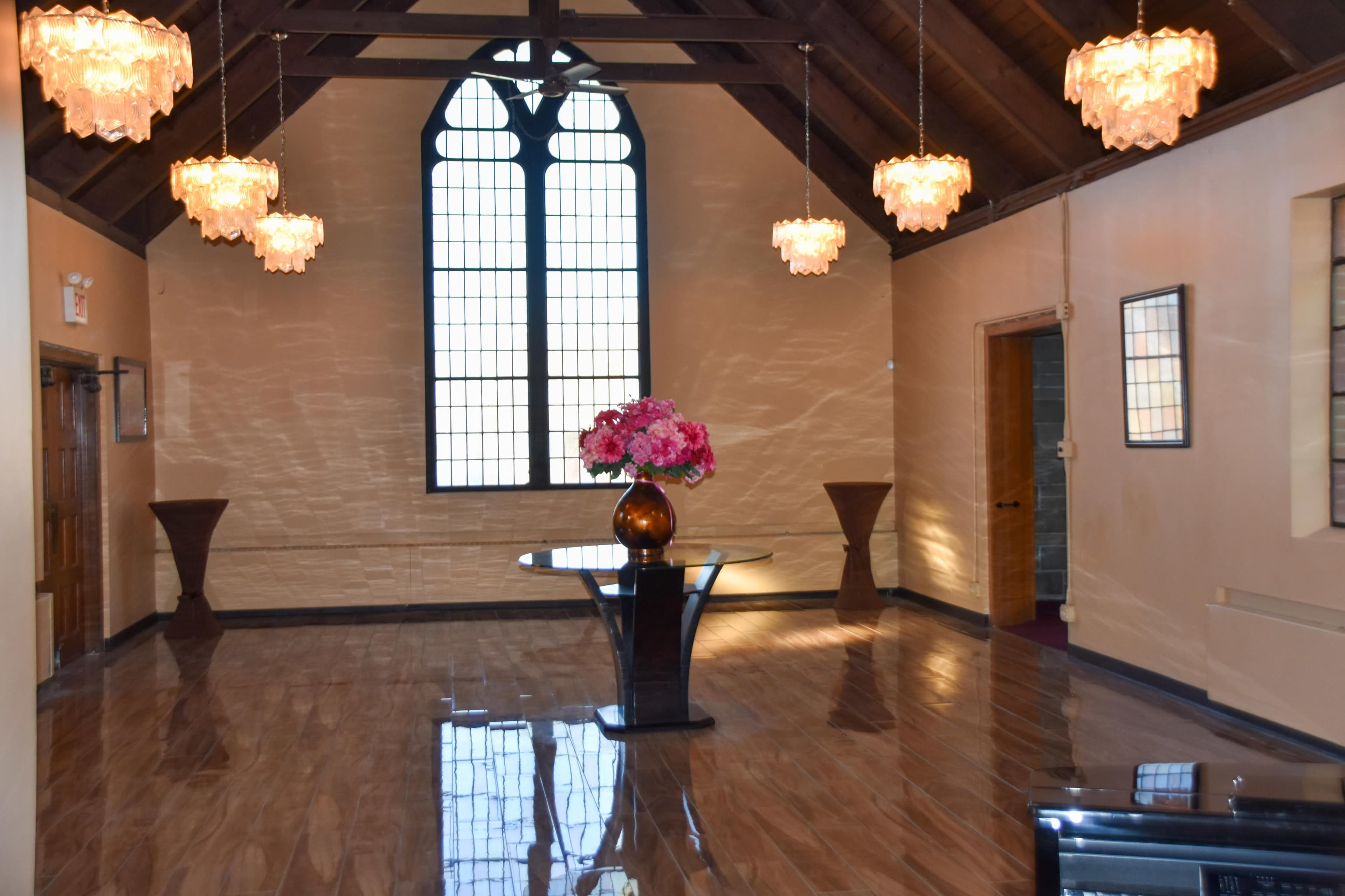 The image shows a spacious, well-lit interior with a high ceiling, featuring a large window and several decorative light fixtures, along with a central table and floral arrangement.