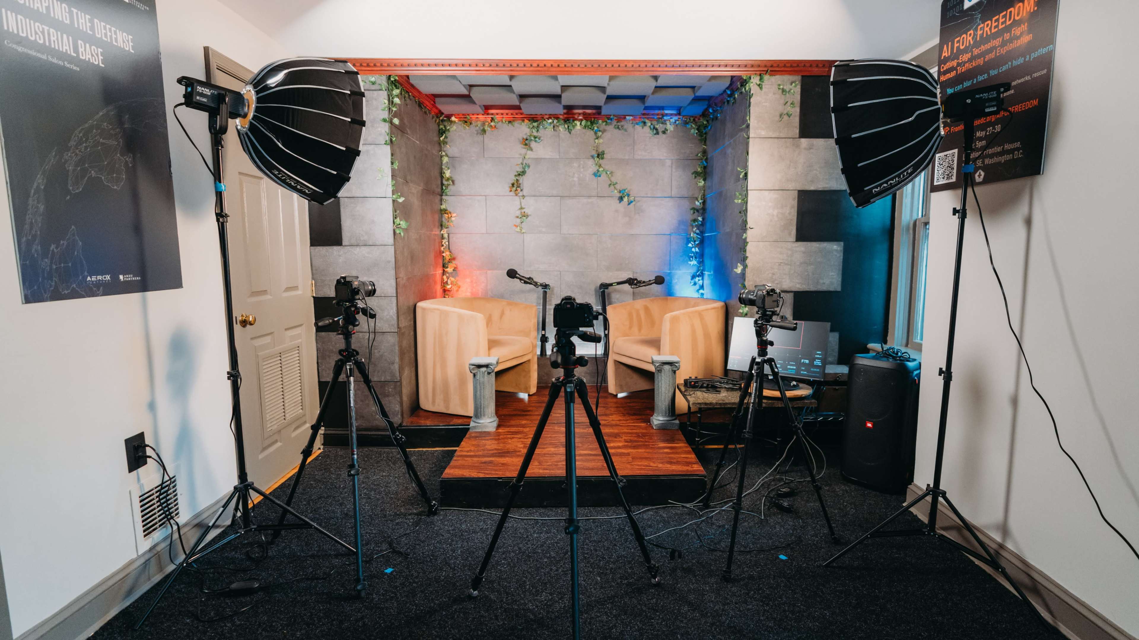 The image shows a podcast recording studio with two chairs, multiple microphone stands, and softbox lights set up against a backdrop of plants and colored lighting.