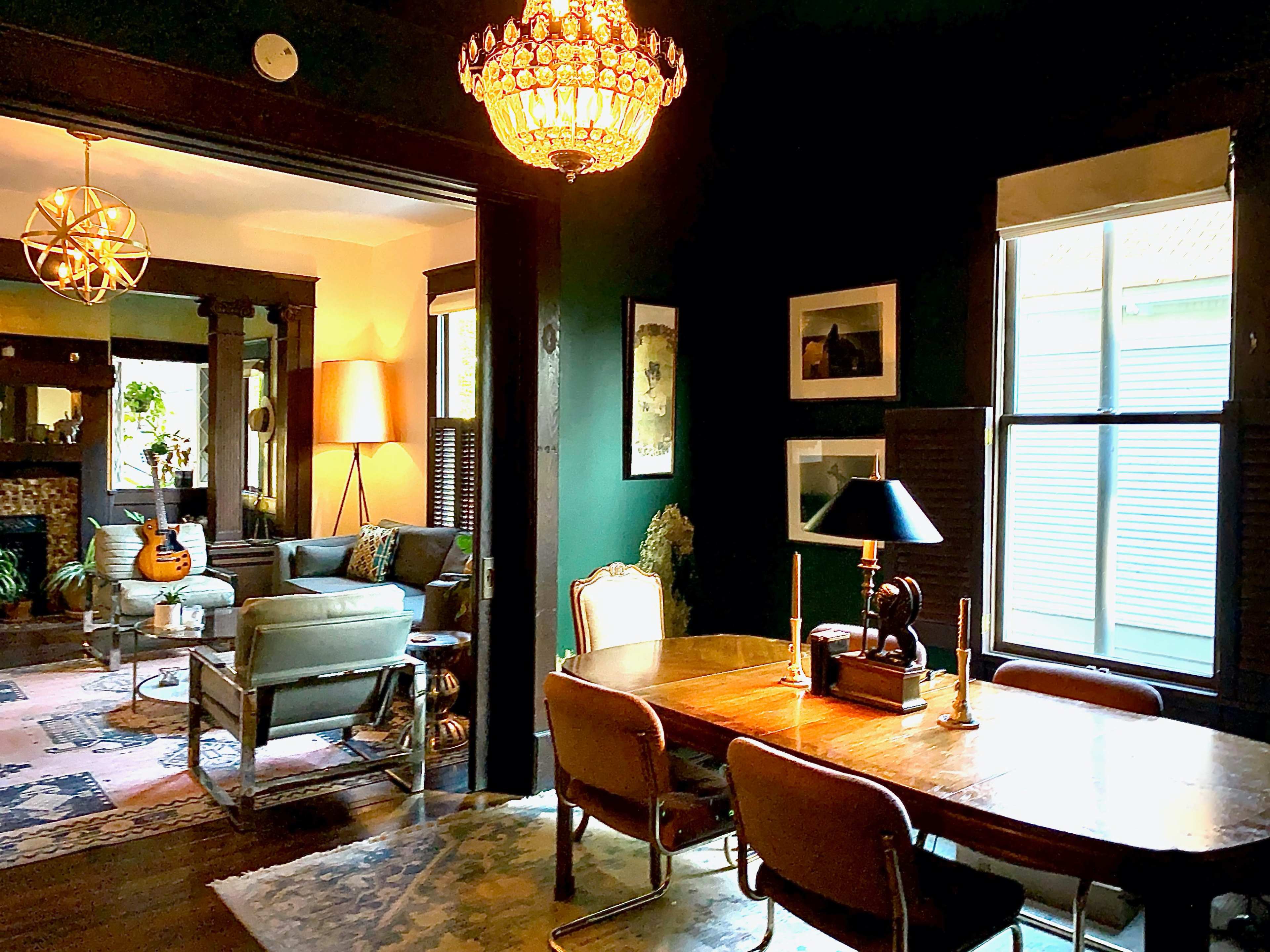 A dining area features a wooden table with chairs, a lamp, and a chandelier, while a living room with a guitar and plant is visible through an open doorway.