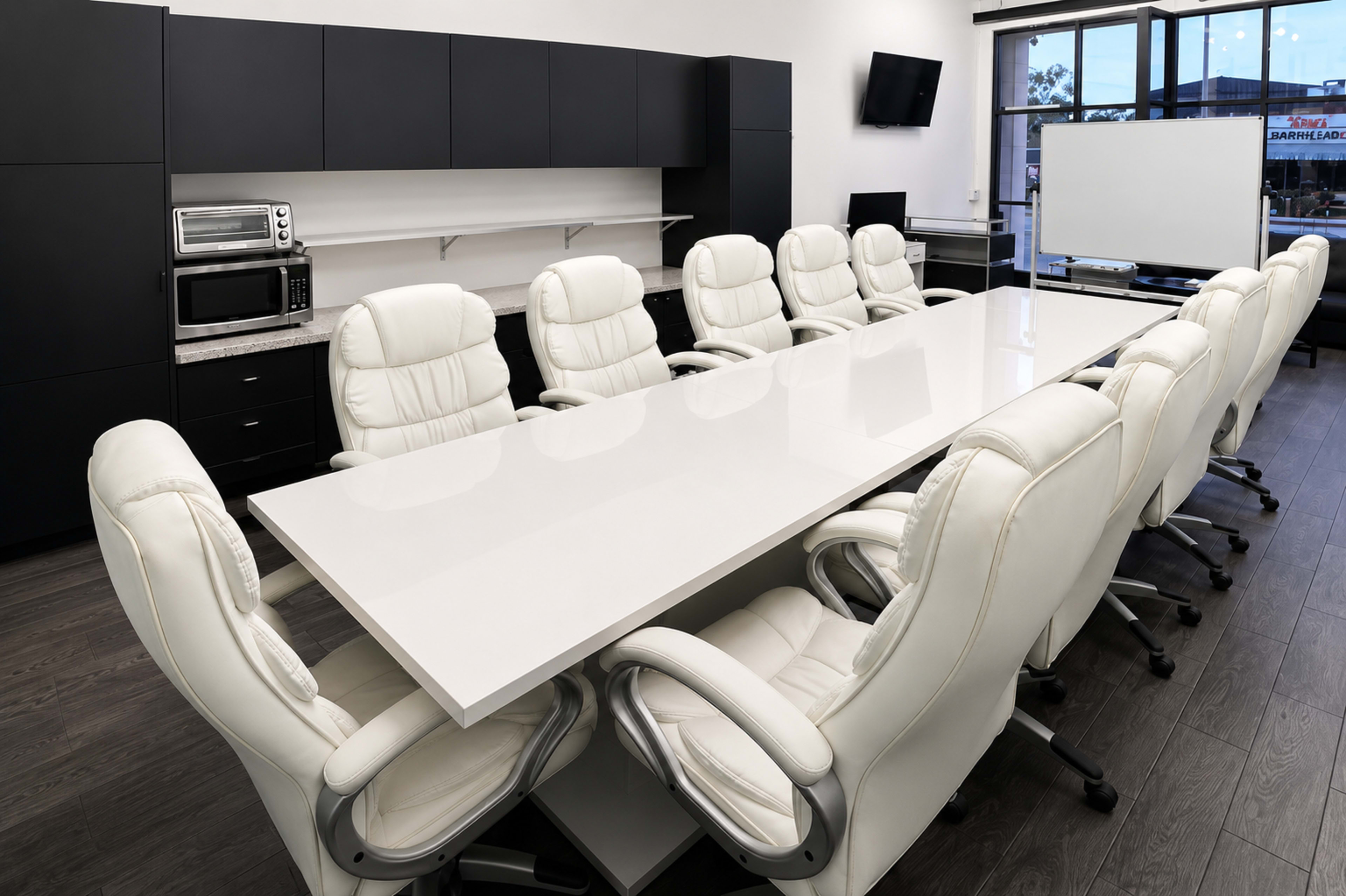 A modern conference room features a long white table surrounded by ten white leather chairs, with a large window and a television mounted on the wall.