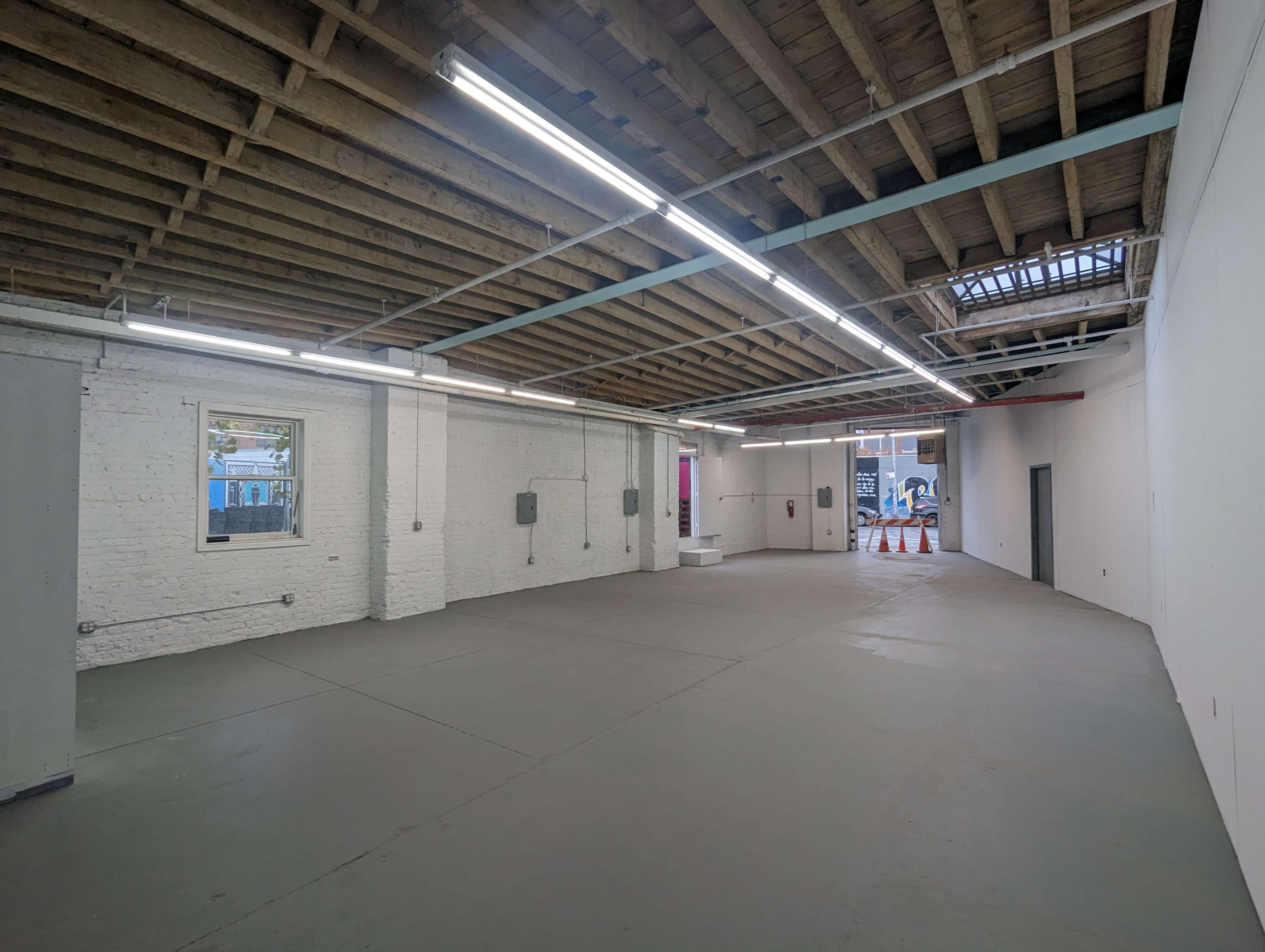 The image shows an empty, industrial-style interior space with a concrete floor, exposed beams, and overhead lighting.