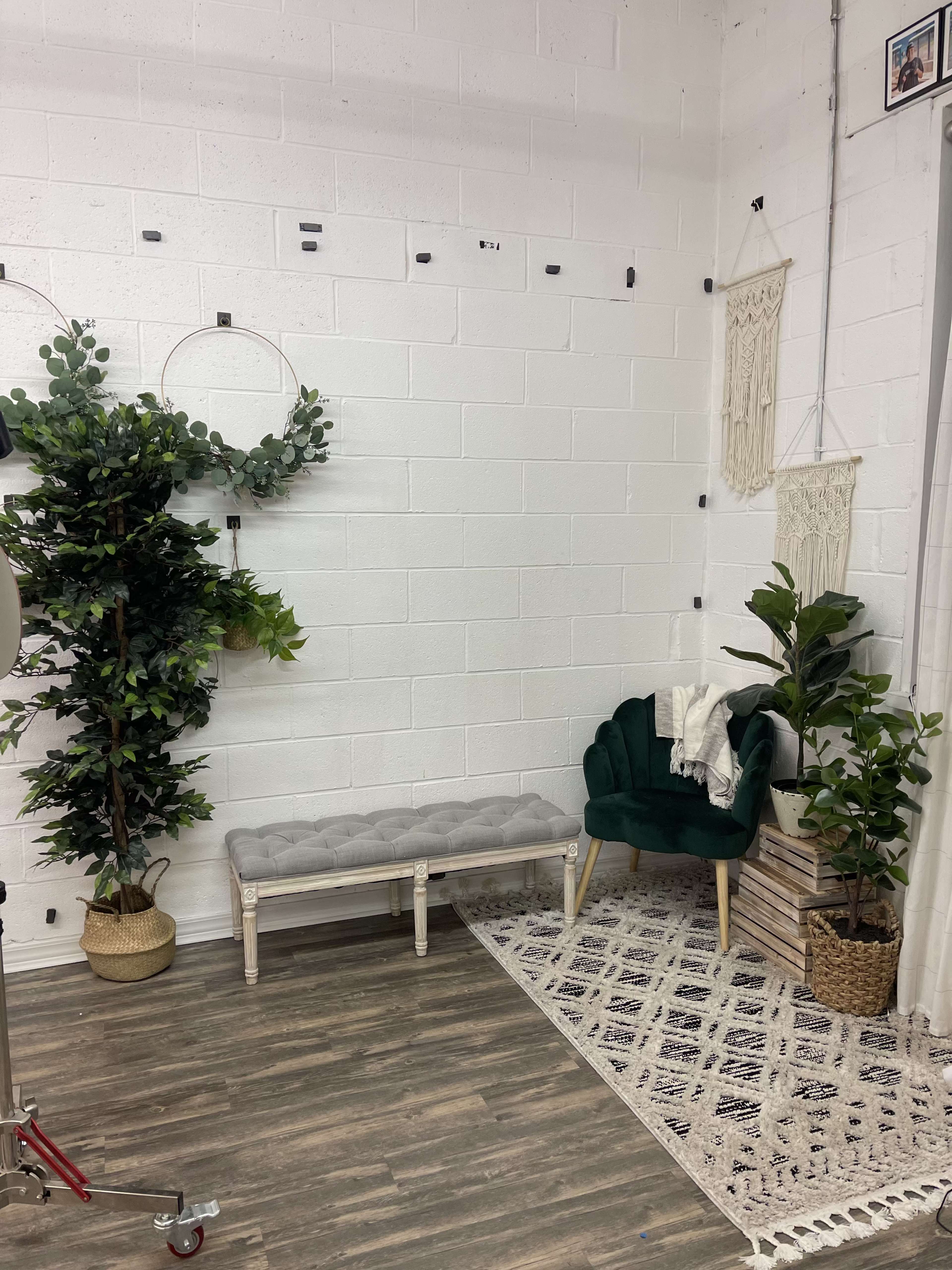 A stylized corner of a room featuring a green upholstered chair, a gray bench, decorative plants, and hanging wall art against a white brick wall.