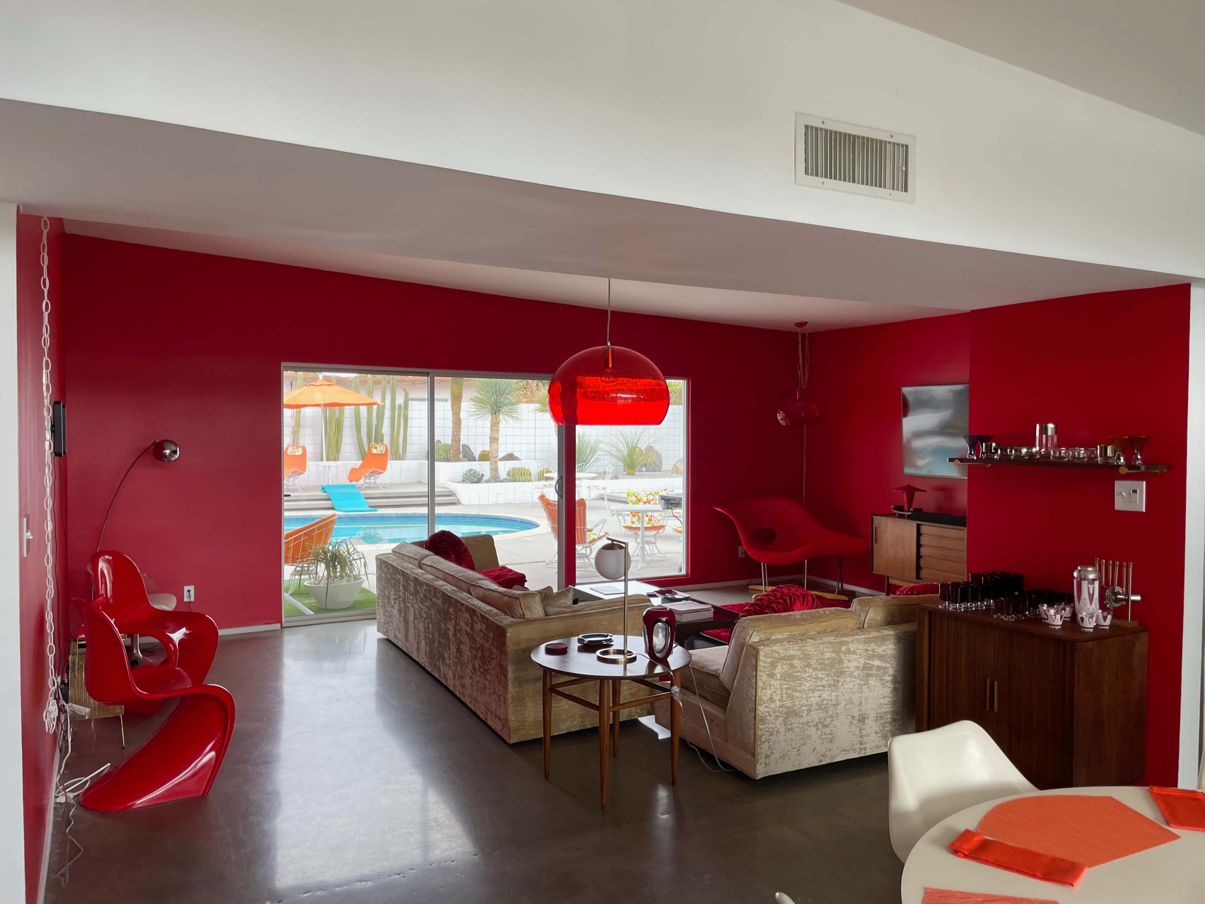 The room features a modern living area with a red accent wall, a beige sofa, and large windows overlooking a poolside area with orange lounge chairs.