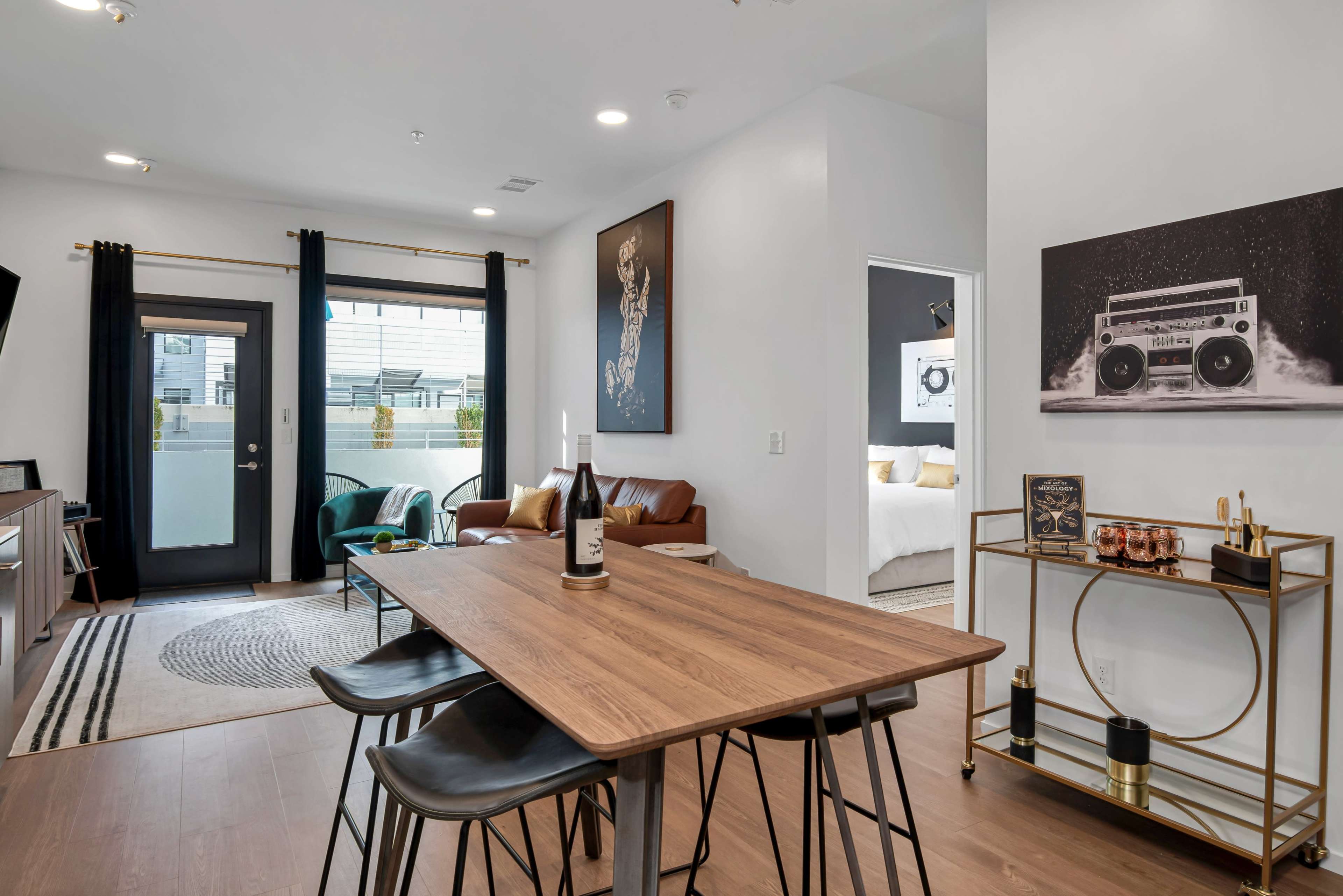 The image shows a modern apartment interior featuring a dining table with chairs, a living area with a brown sofa, and a doorway leading to a balcony.