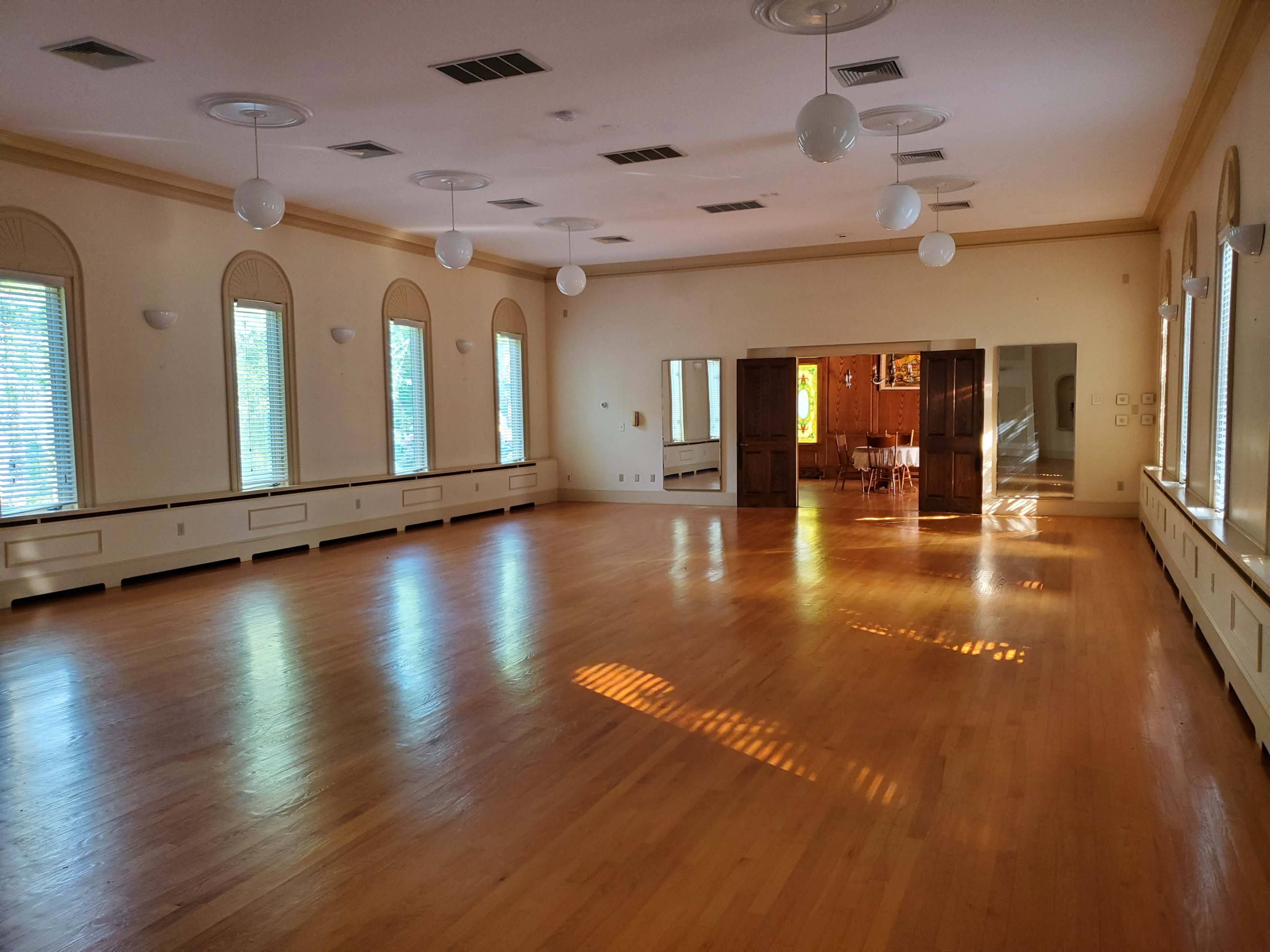 A spacious, well-lit room with hardwood floors, large windows, and a set of mirrors along one wall.