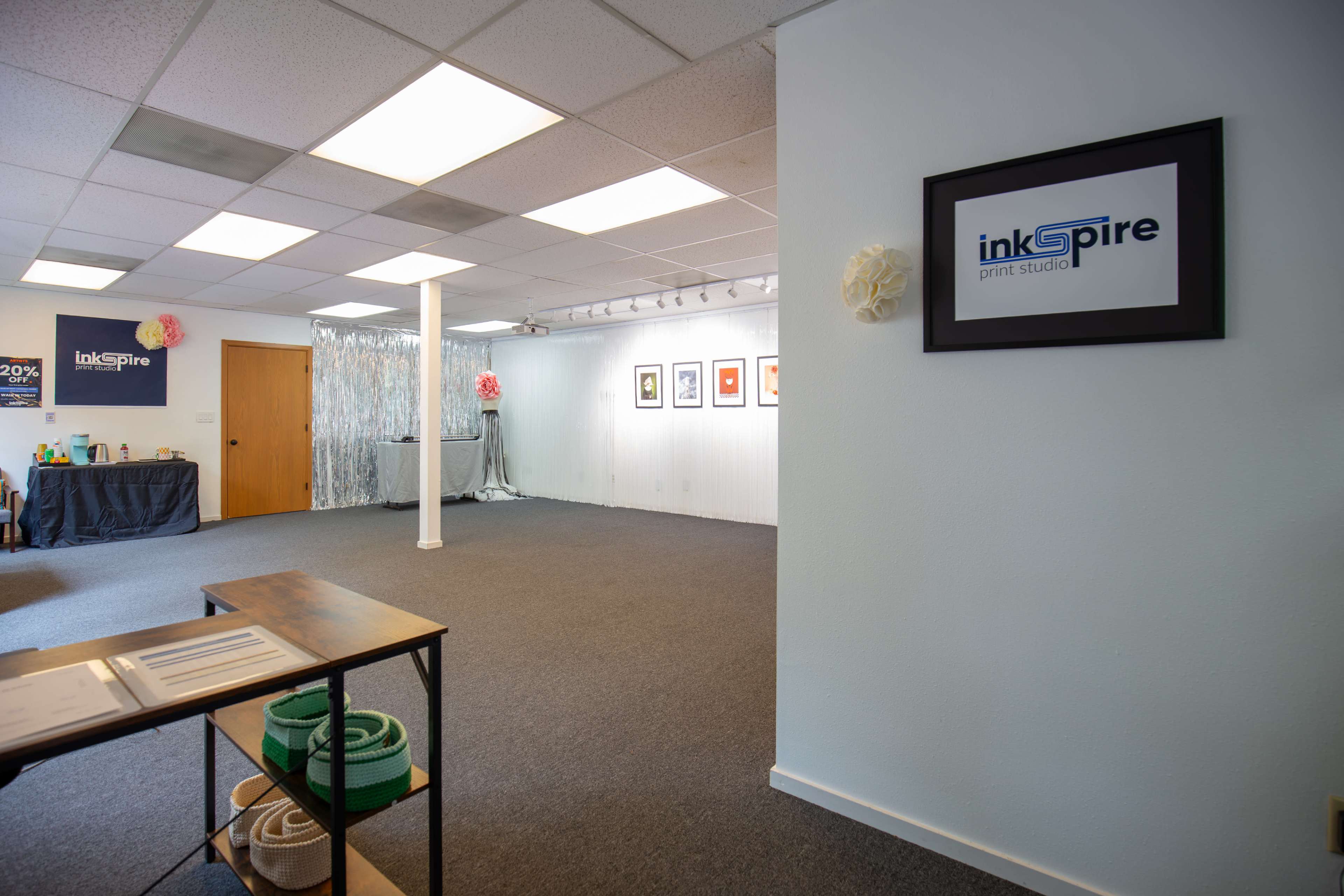 The image shows a print studio with gray carpeting, white walls displaying framed artwork, and a reception area on the left.