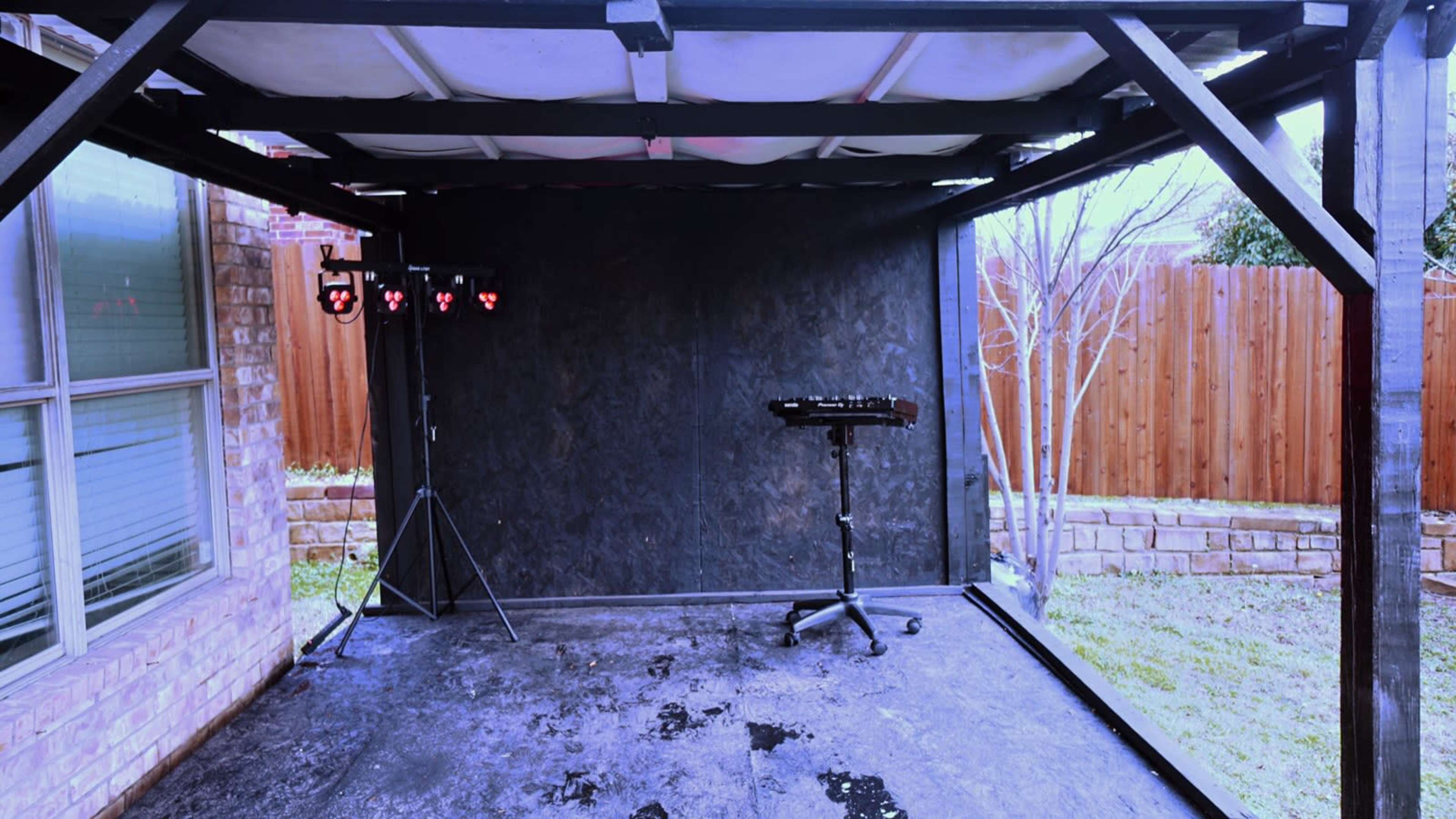 An empty outdoor stage set up with a black backdrop, a lighting rig on a stand, and a keyboard stand, surrounded by a grassy area and a wooden fence.