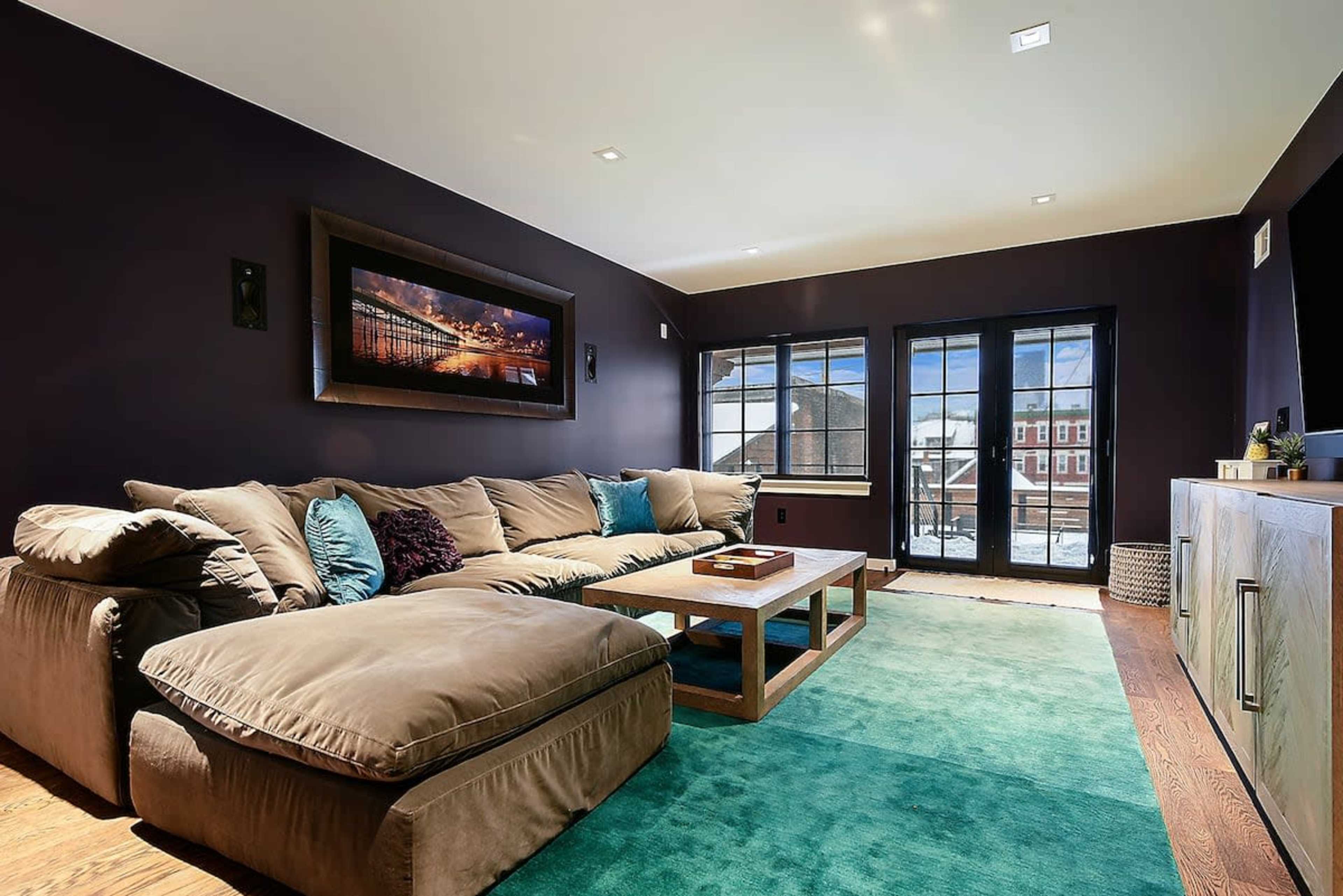A modern living room featuring a large sectional sofa, a wooden coffee table, and a teal area rug, with large windows providing natural light.