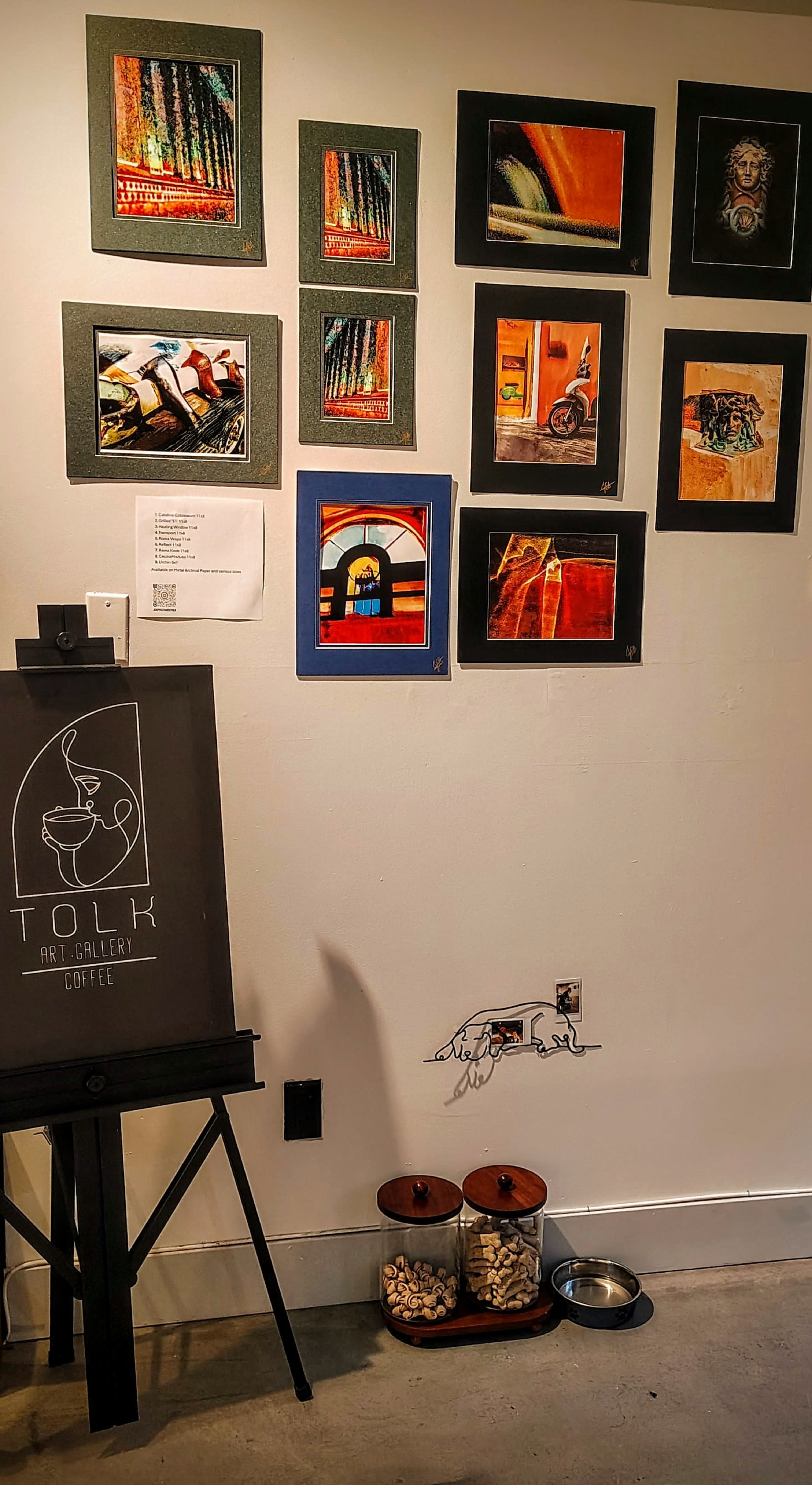 The image shows an art gallery wall featuring various framed artworks alongside a black display sign for "Tolk Art Gallery & Coffee," with a small dog bowl and a glass container of pebbles on the floor.