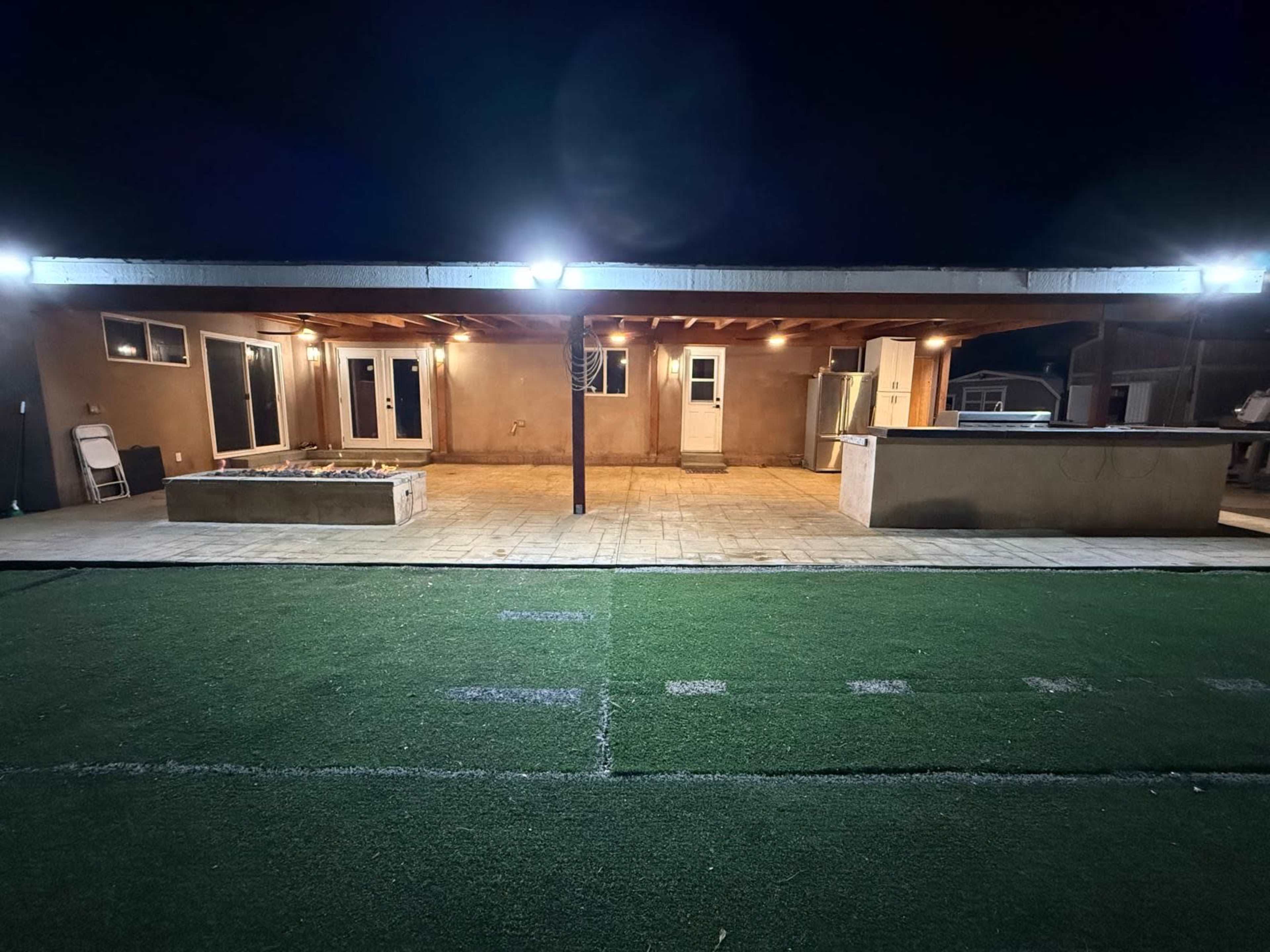 The image shows an outdoor patio area with a covered roof, a fire pit, and an outdoor kitchen, all illuminated by bright lights at night.