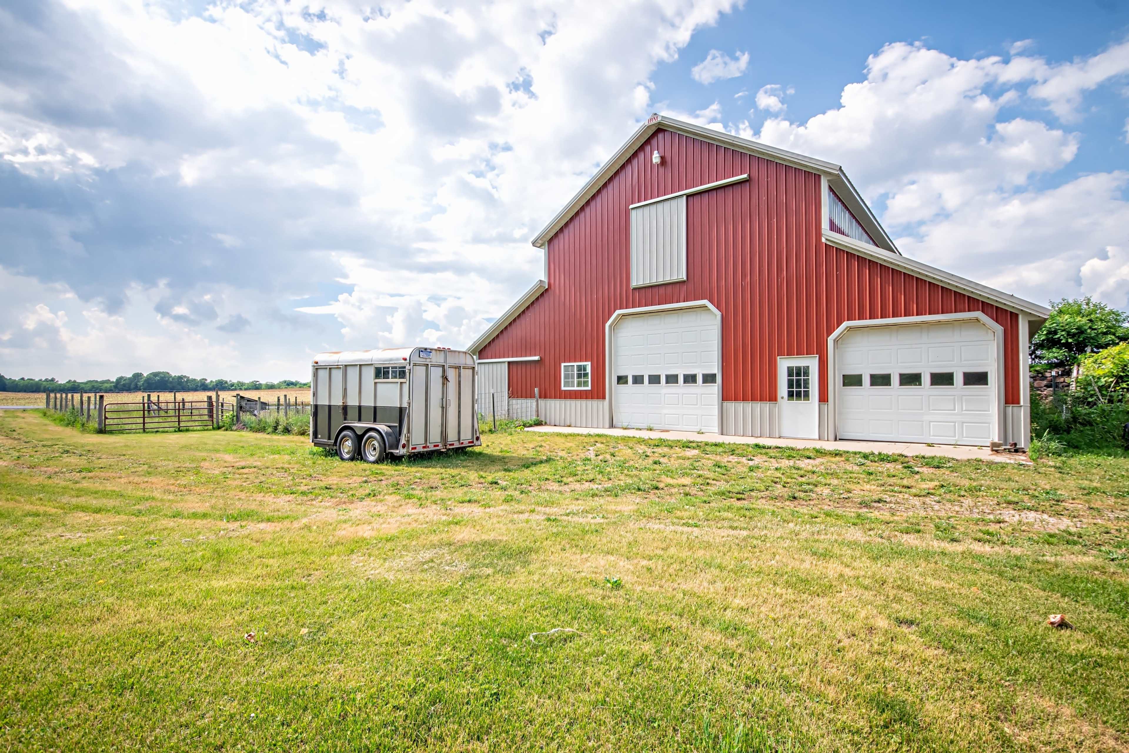 A large red barn with multiple garage doors stands next to a horse trailer on a grassy field under a partly cloudy sky.