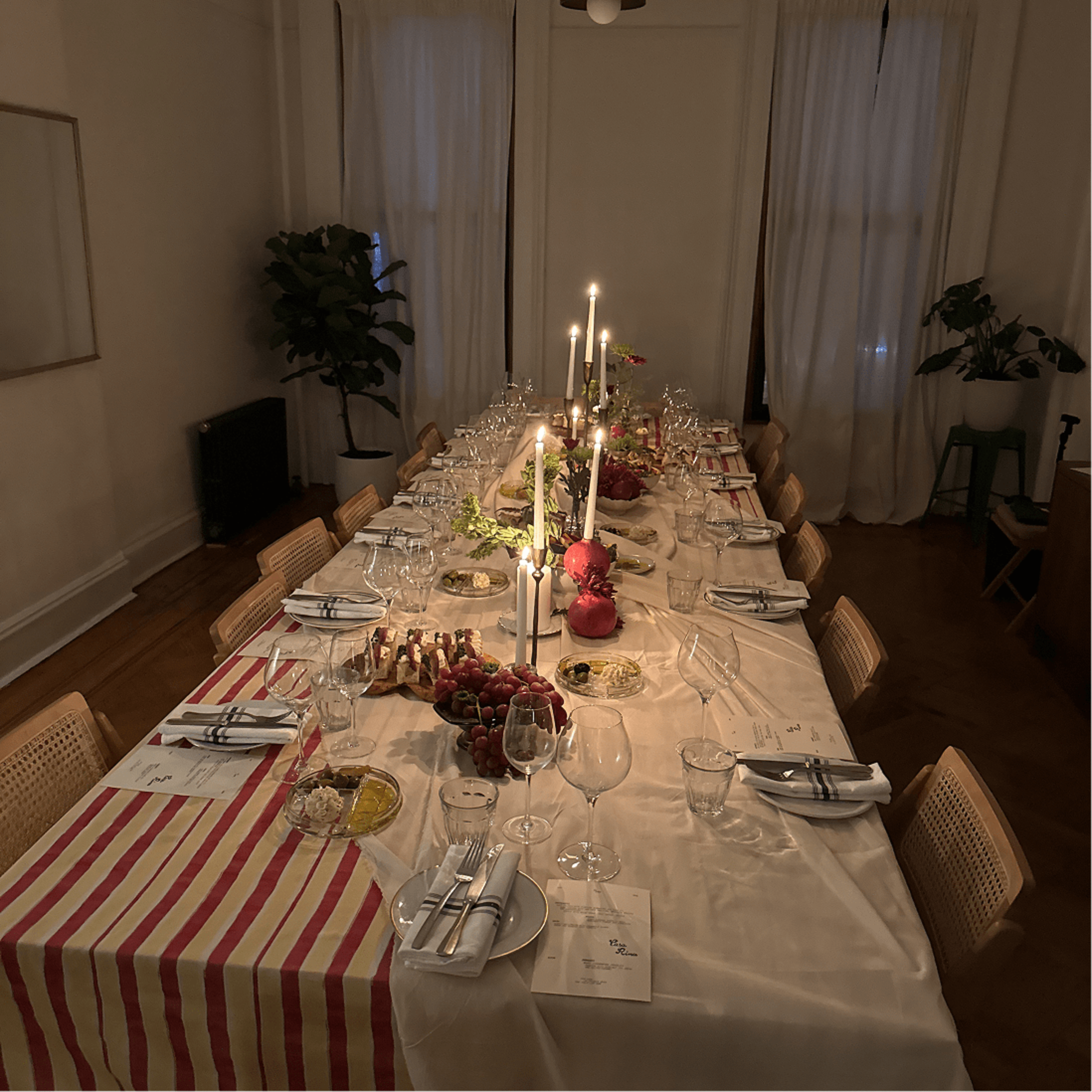 A long dining table is set with candles, glassware, and decorative dishes, illuminated in a softly lit room with sheer curtains.