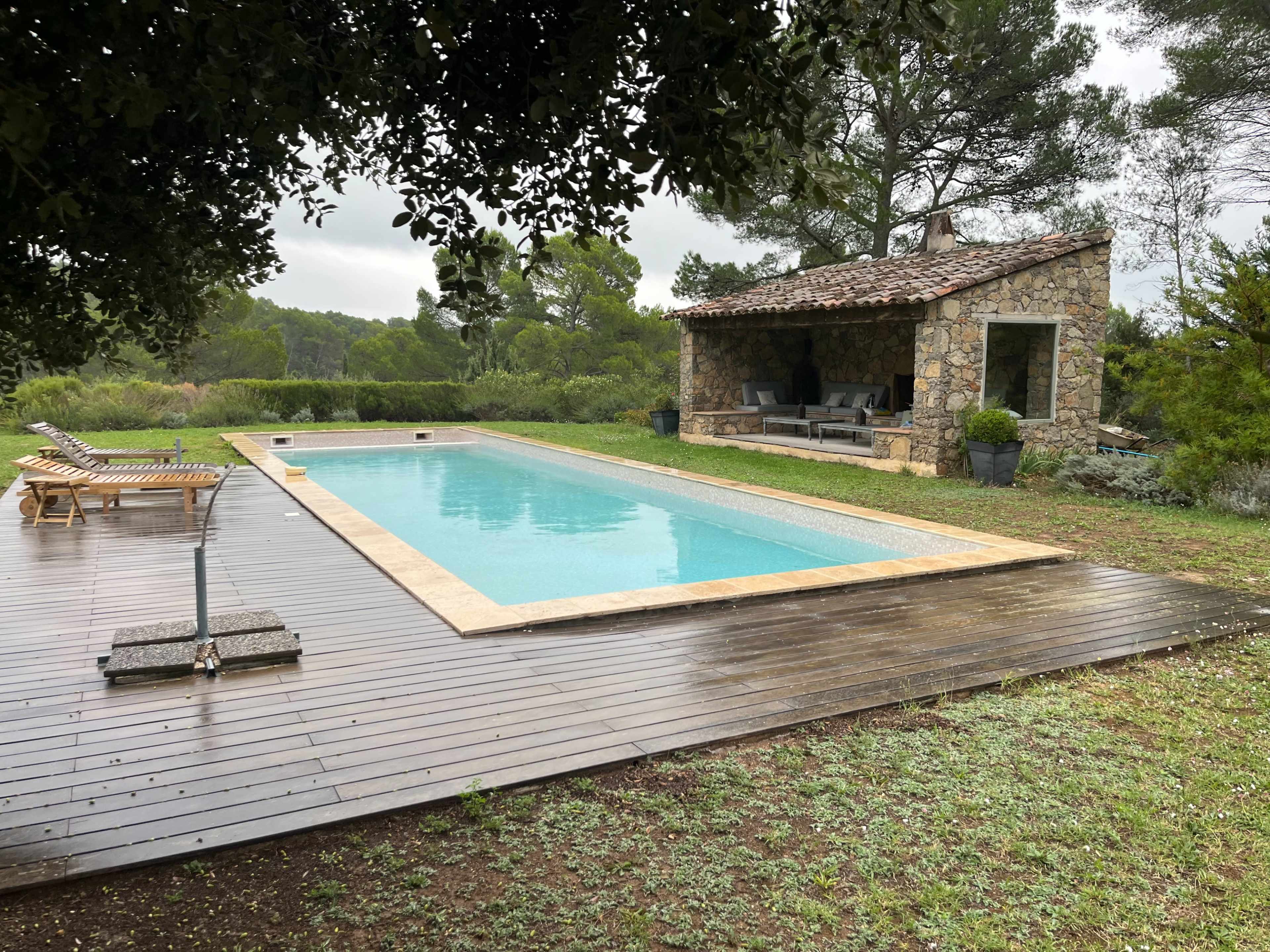 The image shows a rectangular swimming pool surrounded by a wooden deck and a rustic stone building in a natural landscape with trees.