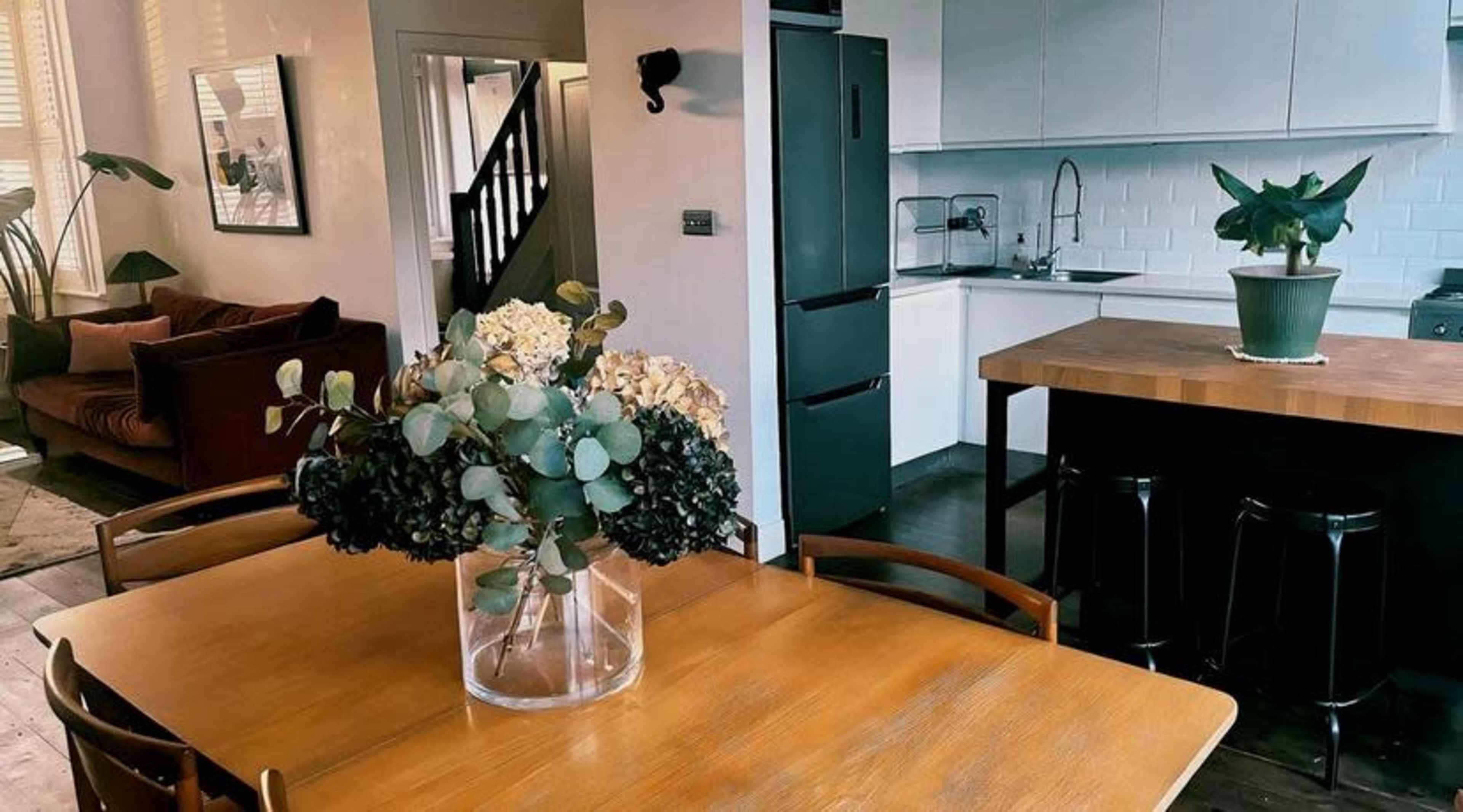 A wooden dining table with a floral centerpiece is set in a modern kitchen and living area featuring a couch and a staircase.