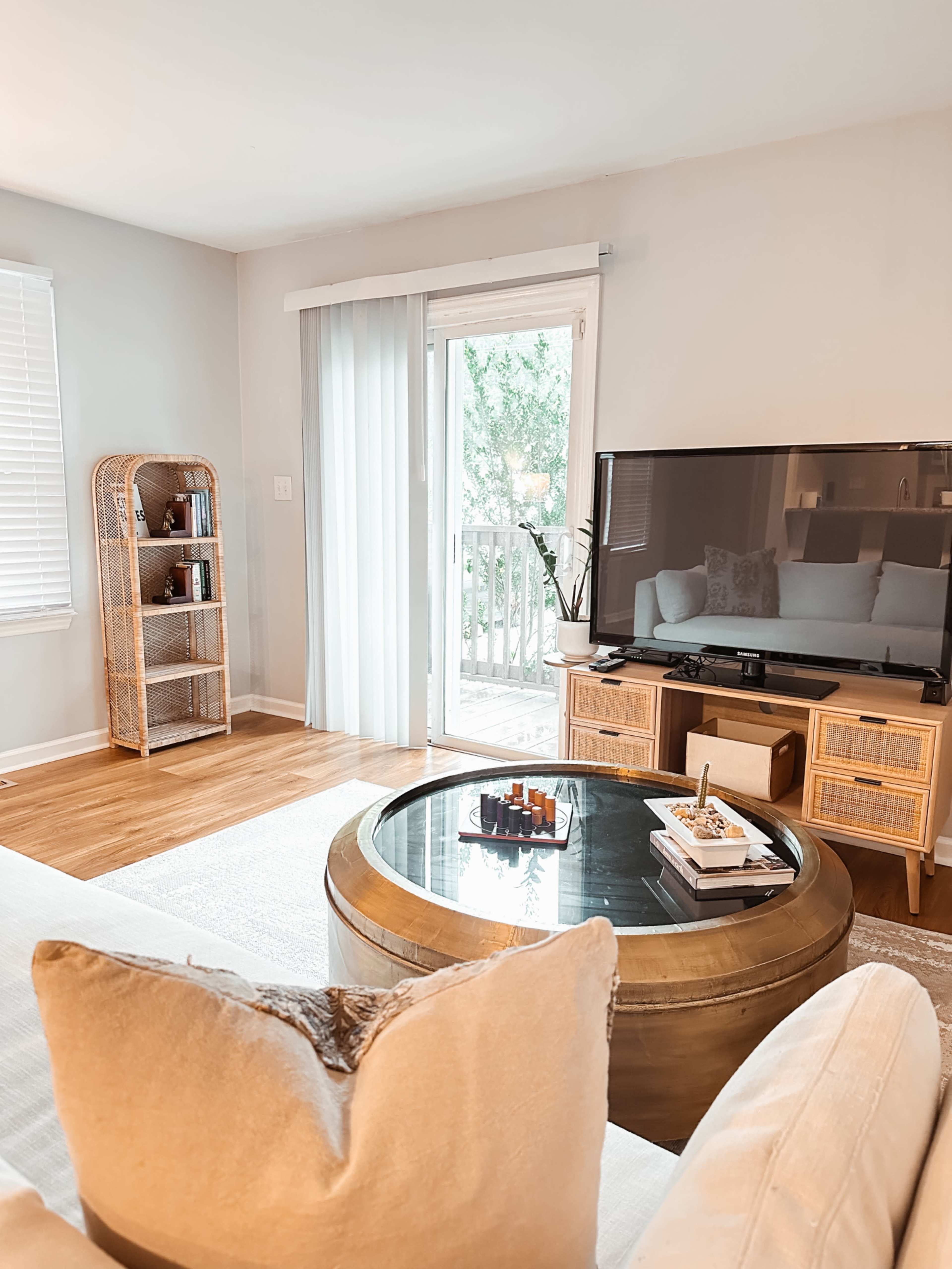 The image shows a bright living room featuring a glass coffee table, a television stand, and a sliding door leading to a patio.