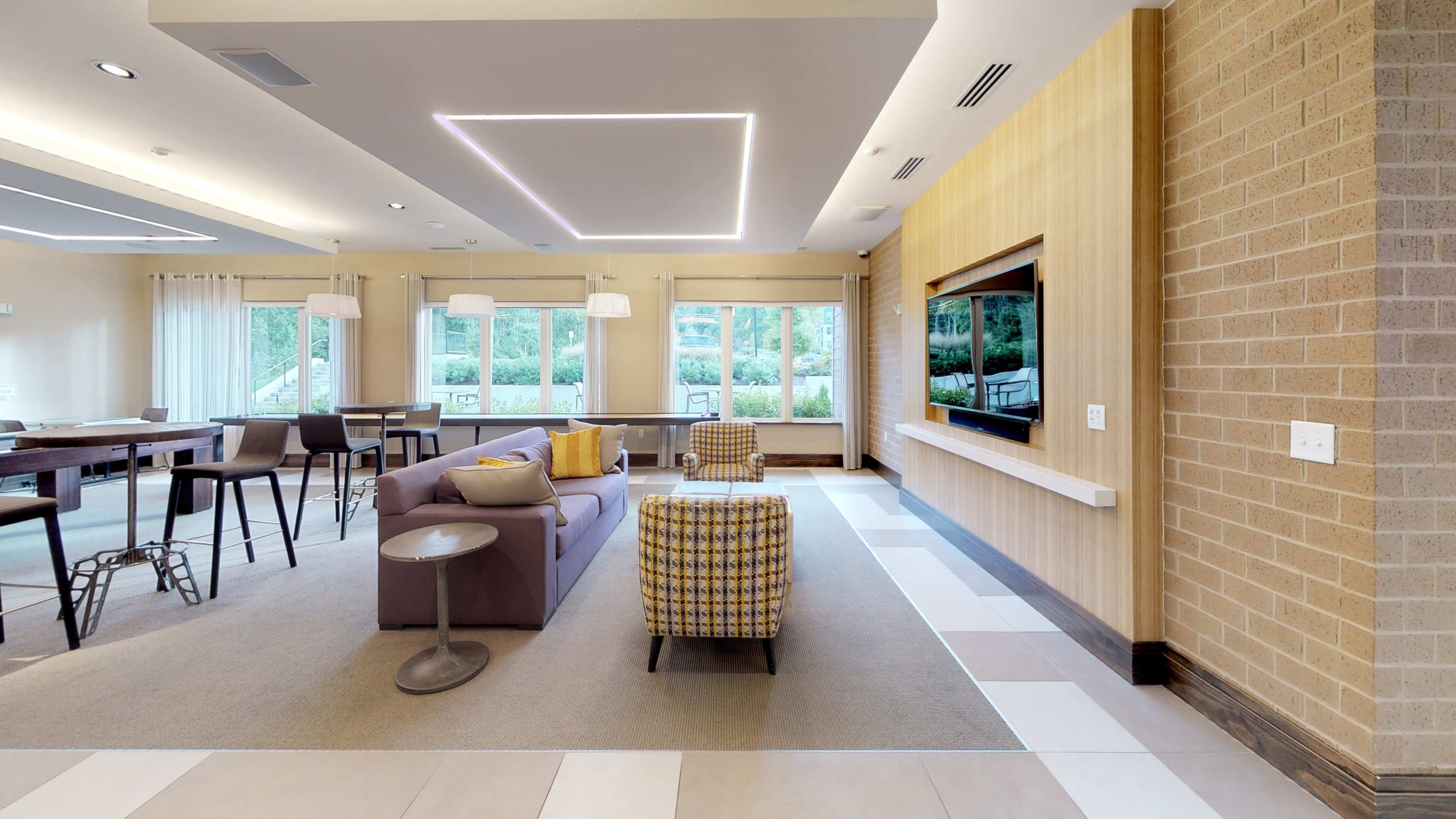 A modern lounge area featuring a purple sofa, a checkered ottoman, a flat-screen TV on a wall, and large windows that provide natural light.