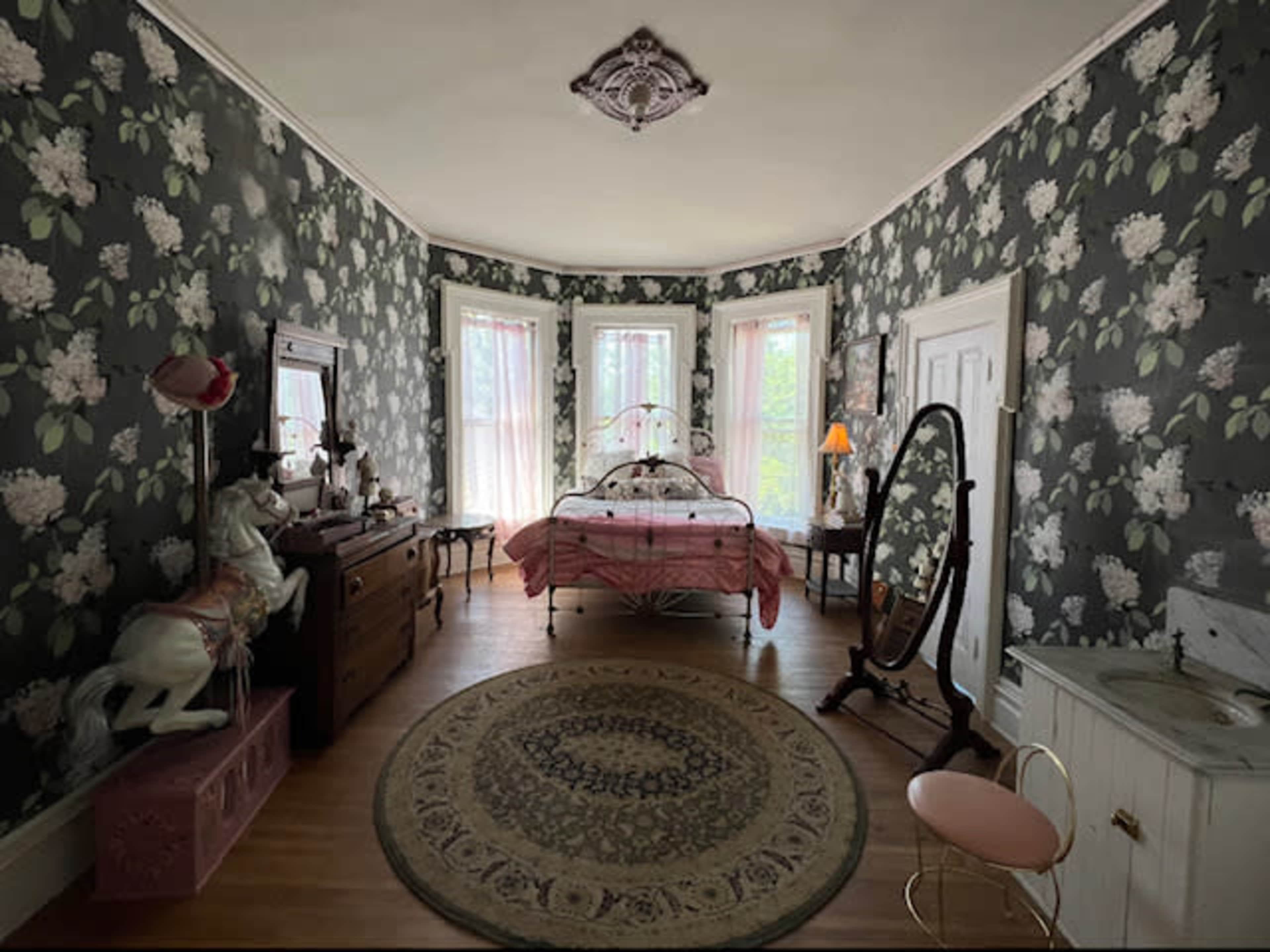 A vintage bedroom featuring floral wallpaper, a bed with a patterned cover, a vanity mirror, and a decorative rug in the center.