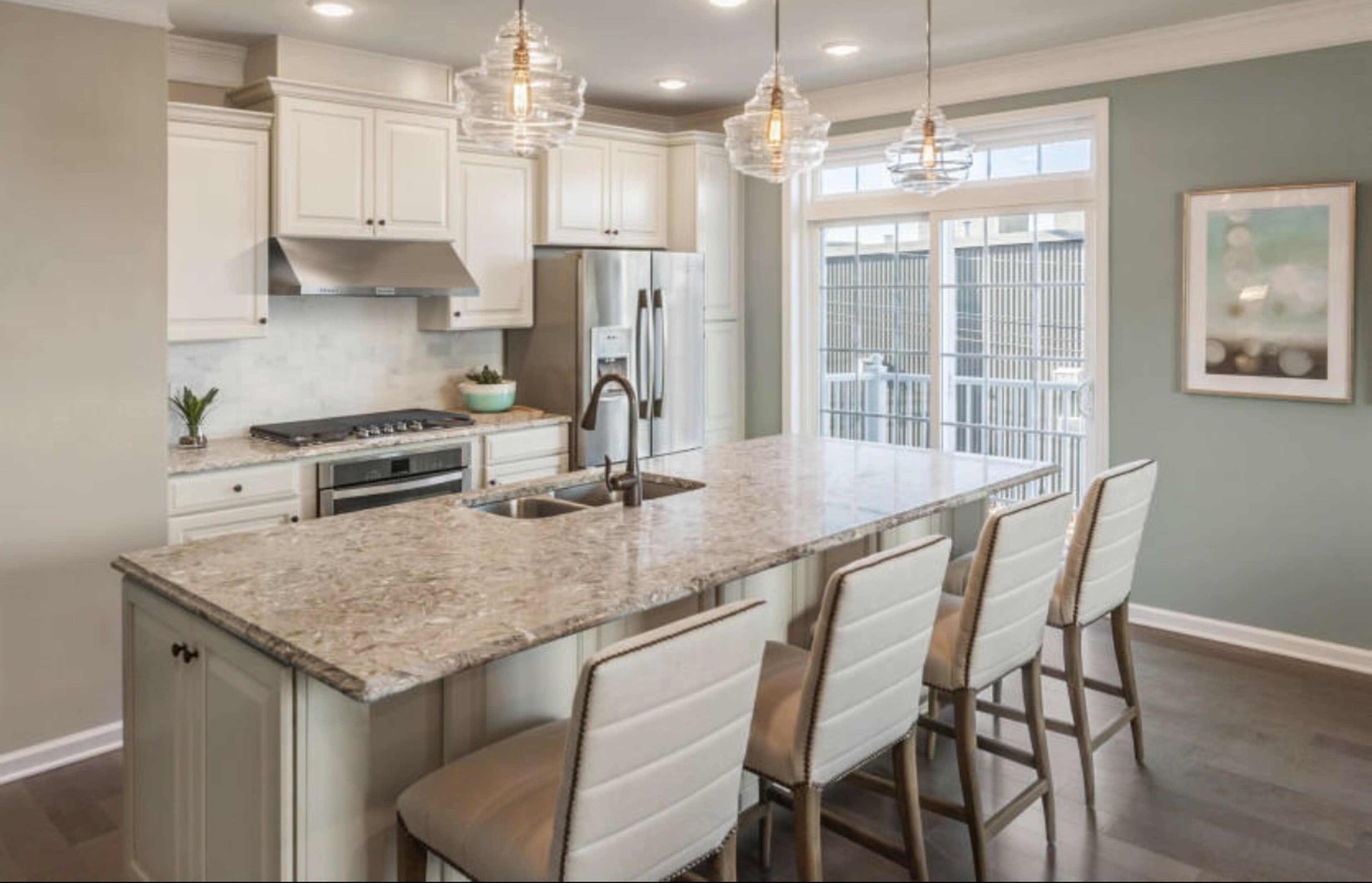 A modern kitchen with a large granite island, six beige bar stools, and stainless steel appliances.