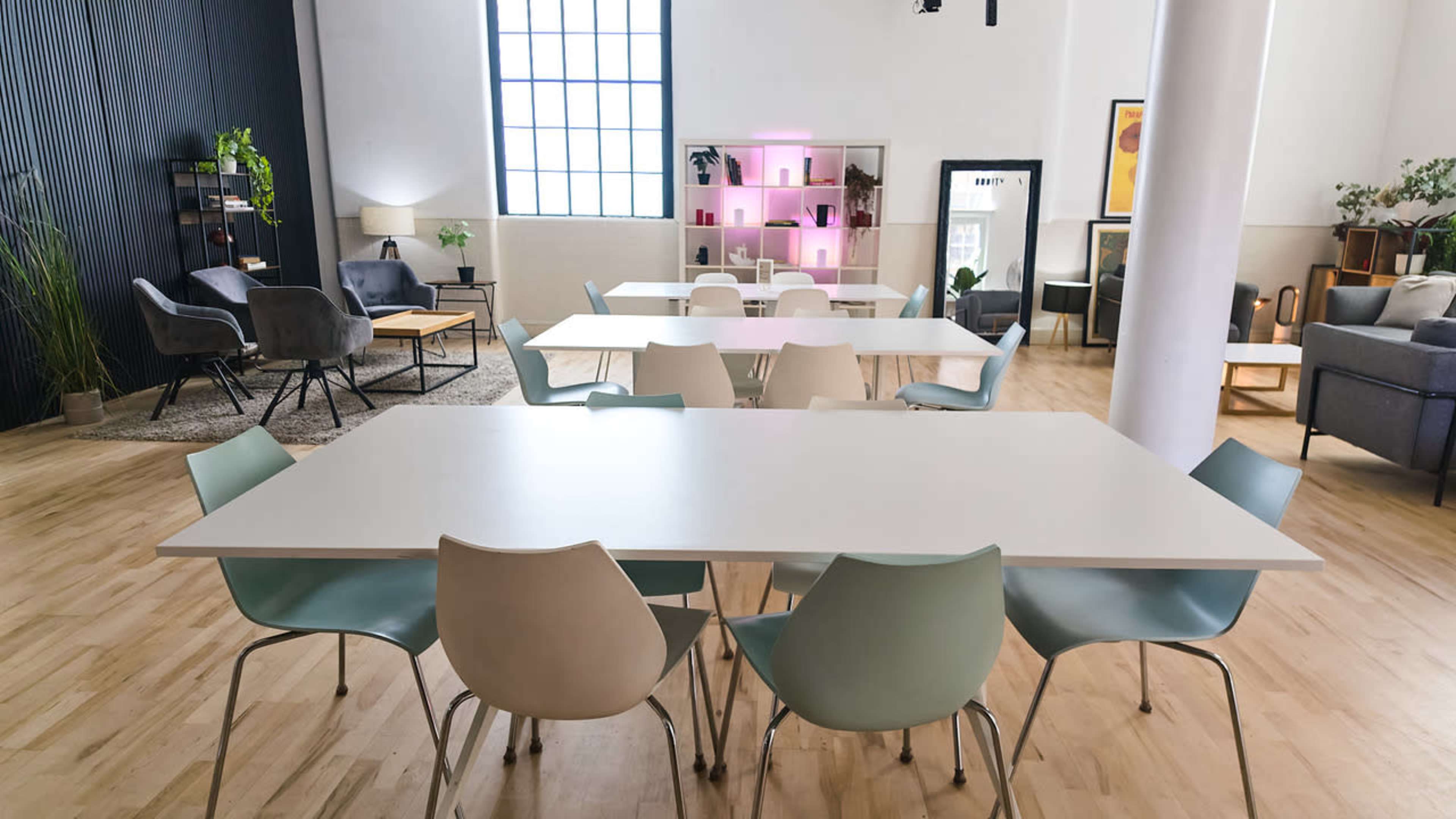 The image shows a spacious and well-lit room filled with several tables and chairs arranged for seating, alongside comfortable lounge areas with plants and decorative shelves.