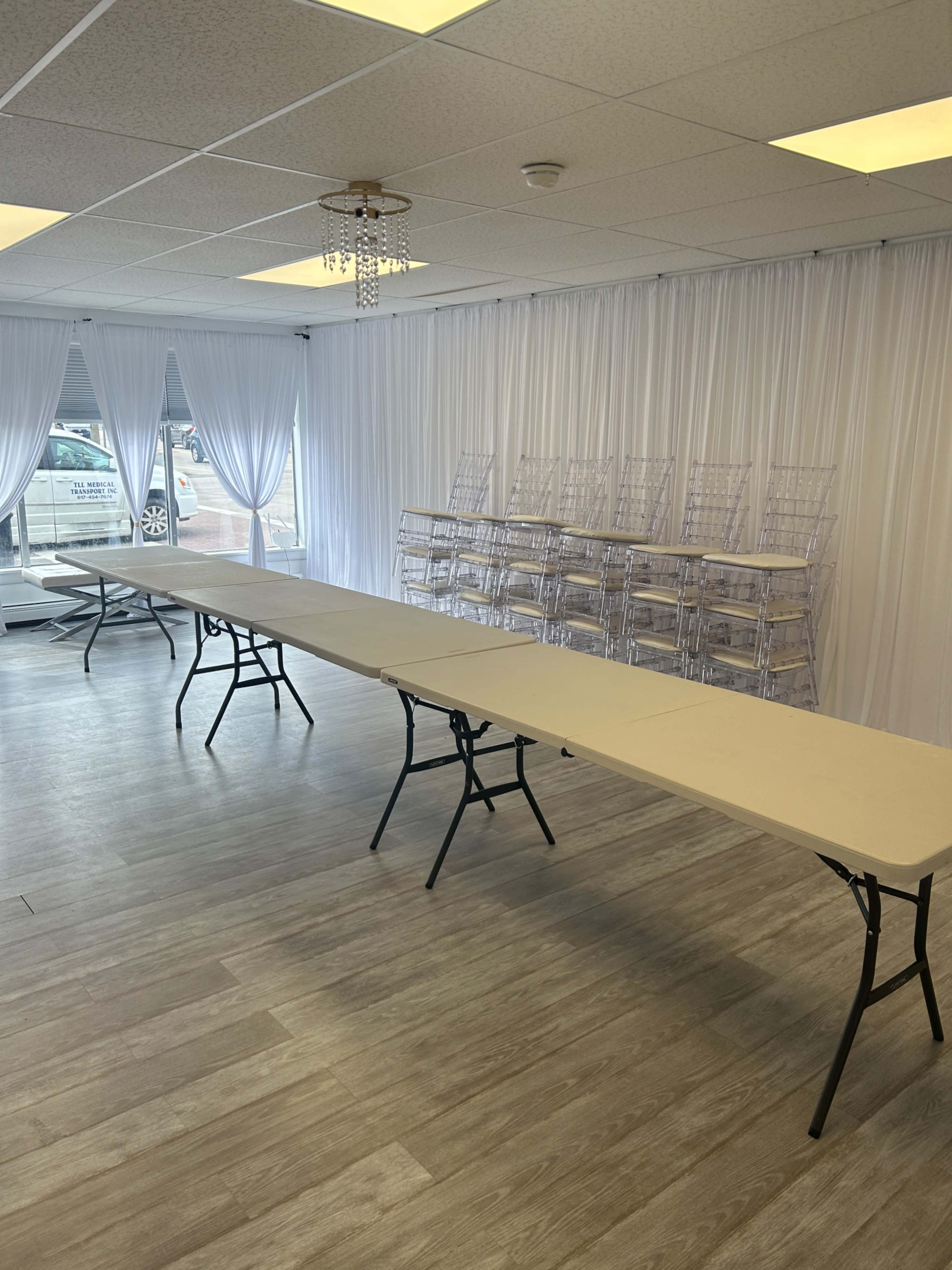 The image shows a spacious indoor venue with a long, empty table set up in the center and multiple clear acrylic chairs stacked in the background.