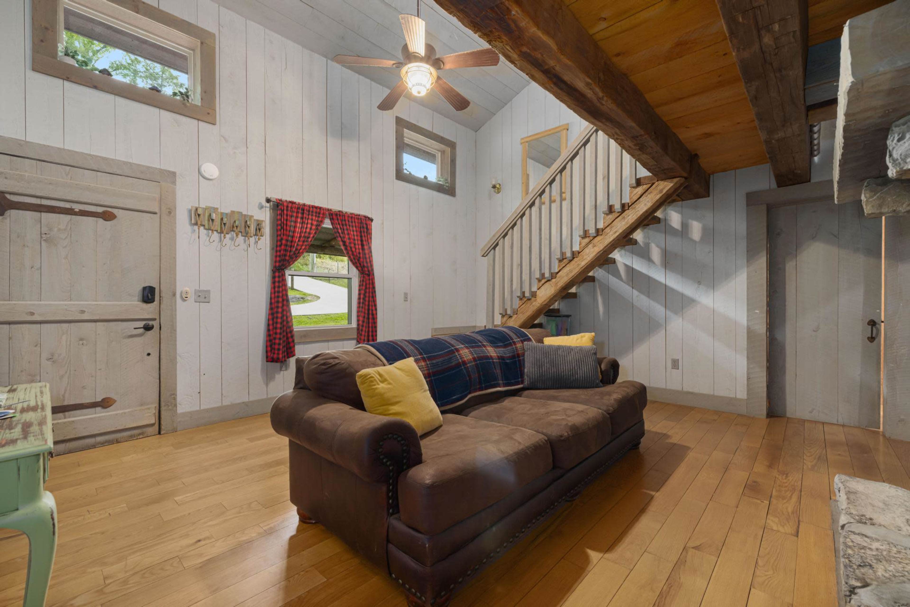 The image shows a cozy living room featuring a brown sofa with yellow pillows, wooden flooring, and a staircase leading to an upper level, along with a large window and a door in the background.