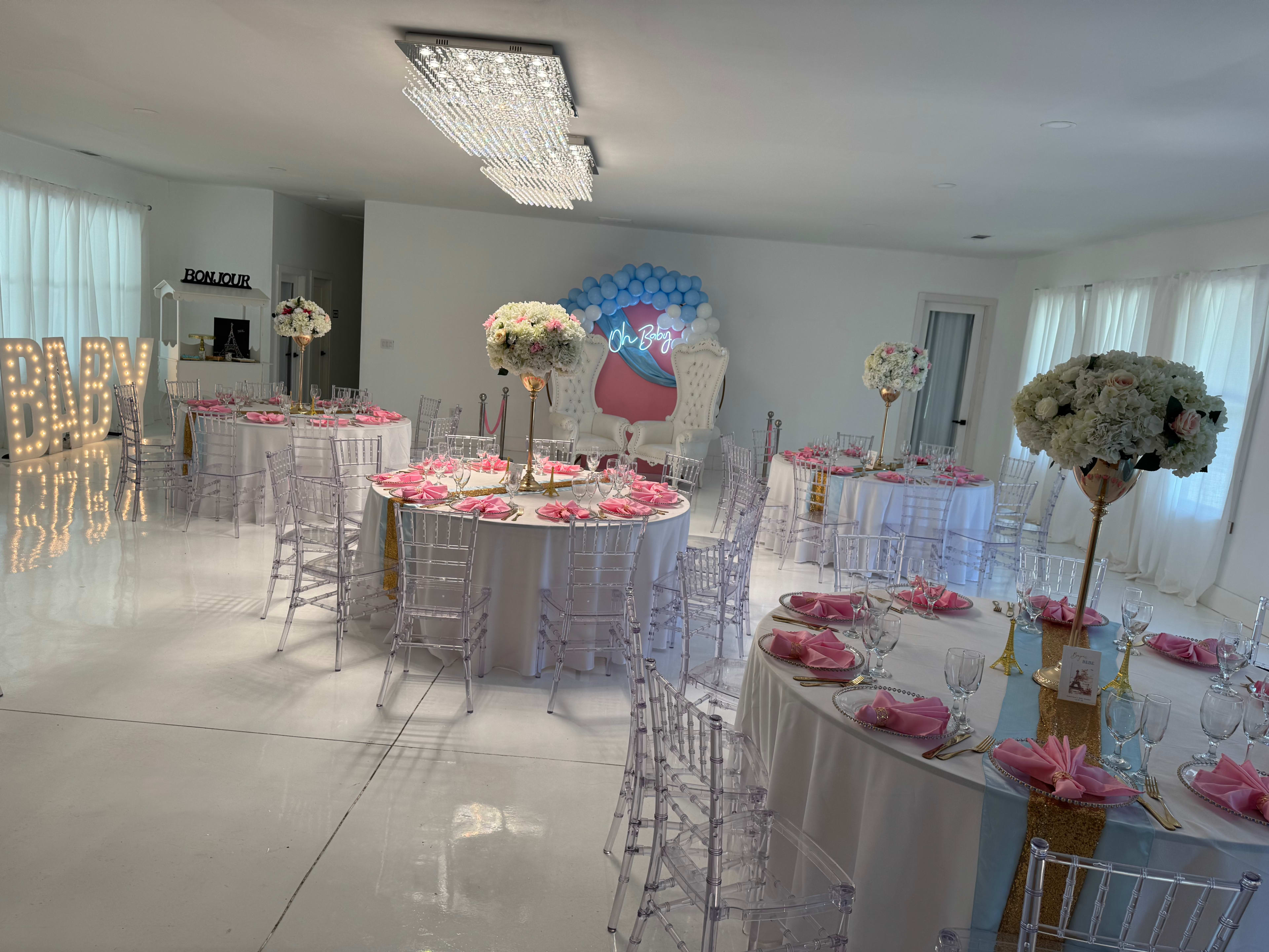 The image shows a well-decorated event space with round tables set for a celebration, featuring pink table settings, floral centerpieces, and a large backdrop with the text "Oh Baby."