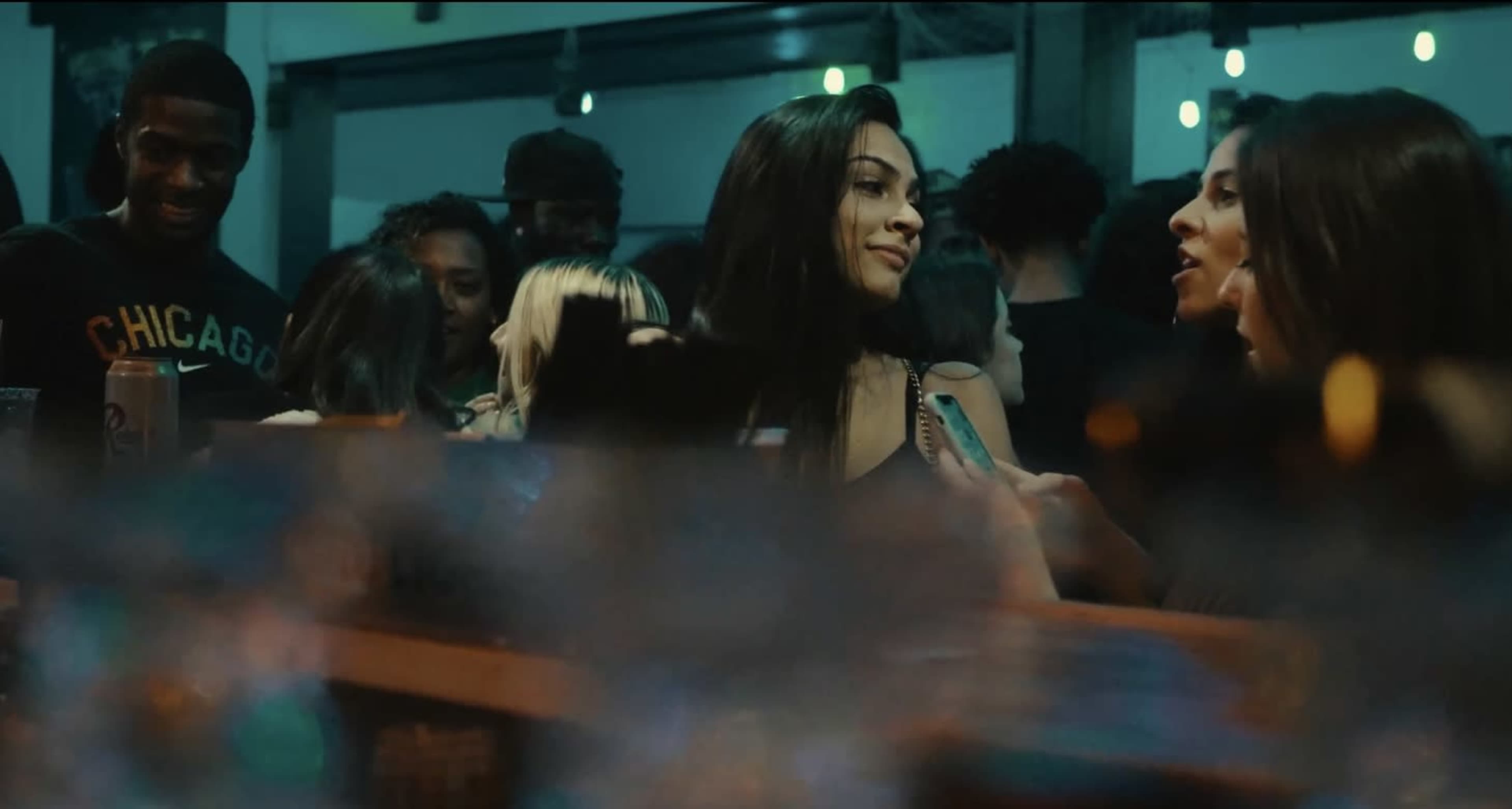 A lively bar scene features a crowd of people with two women engaging in conversation near the foreground.