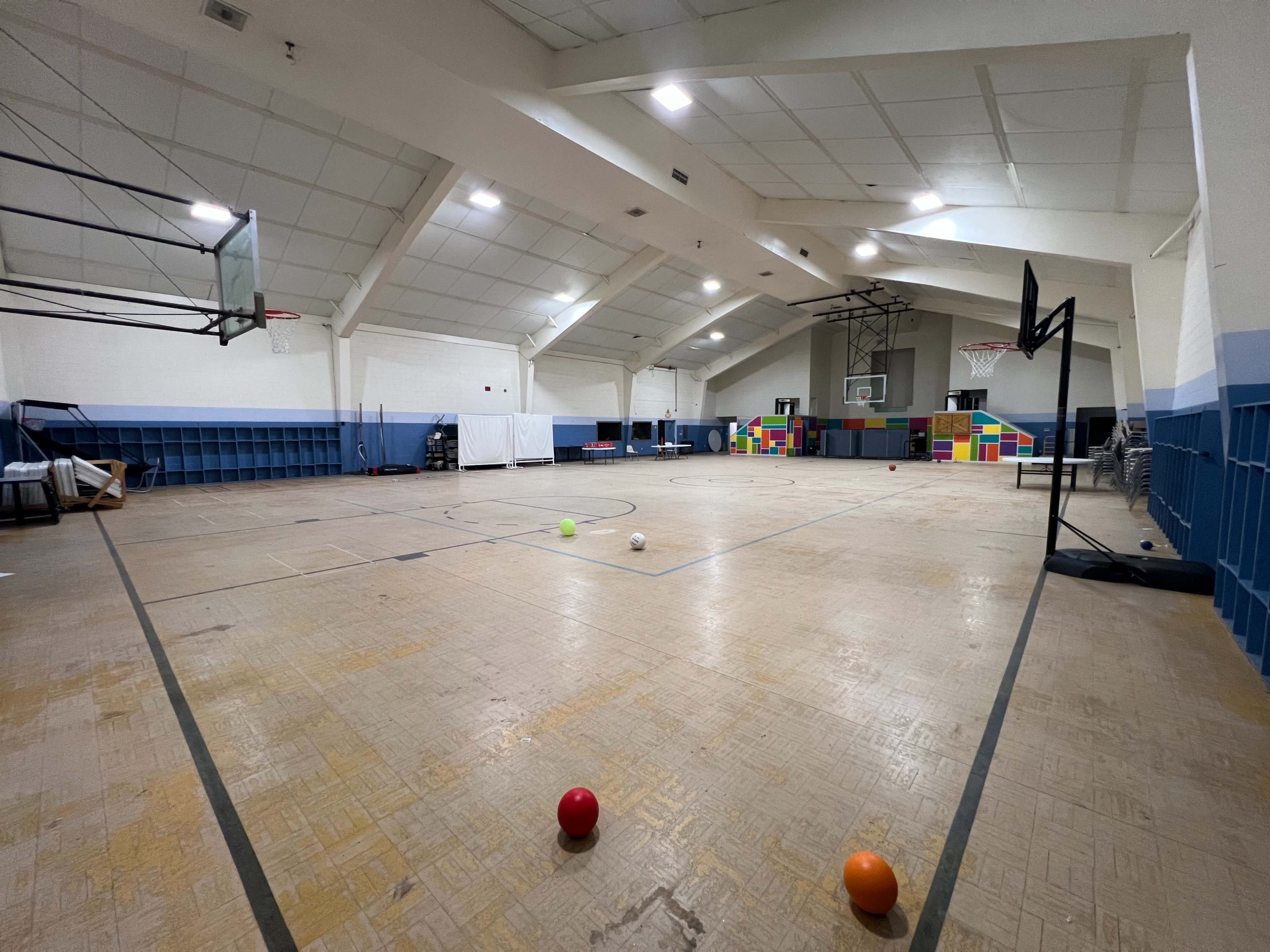 The interior of a gymnasium features a wooden floor, basketball hoops, and colorful play equipment scattered throughout the space.