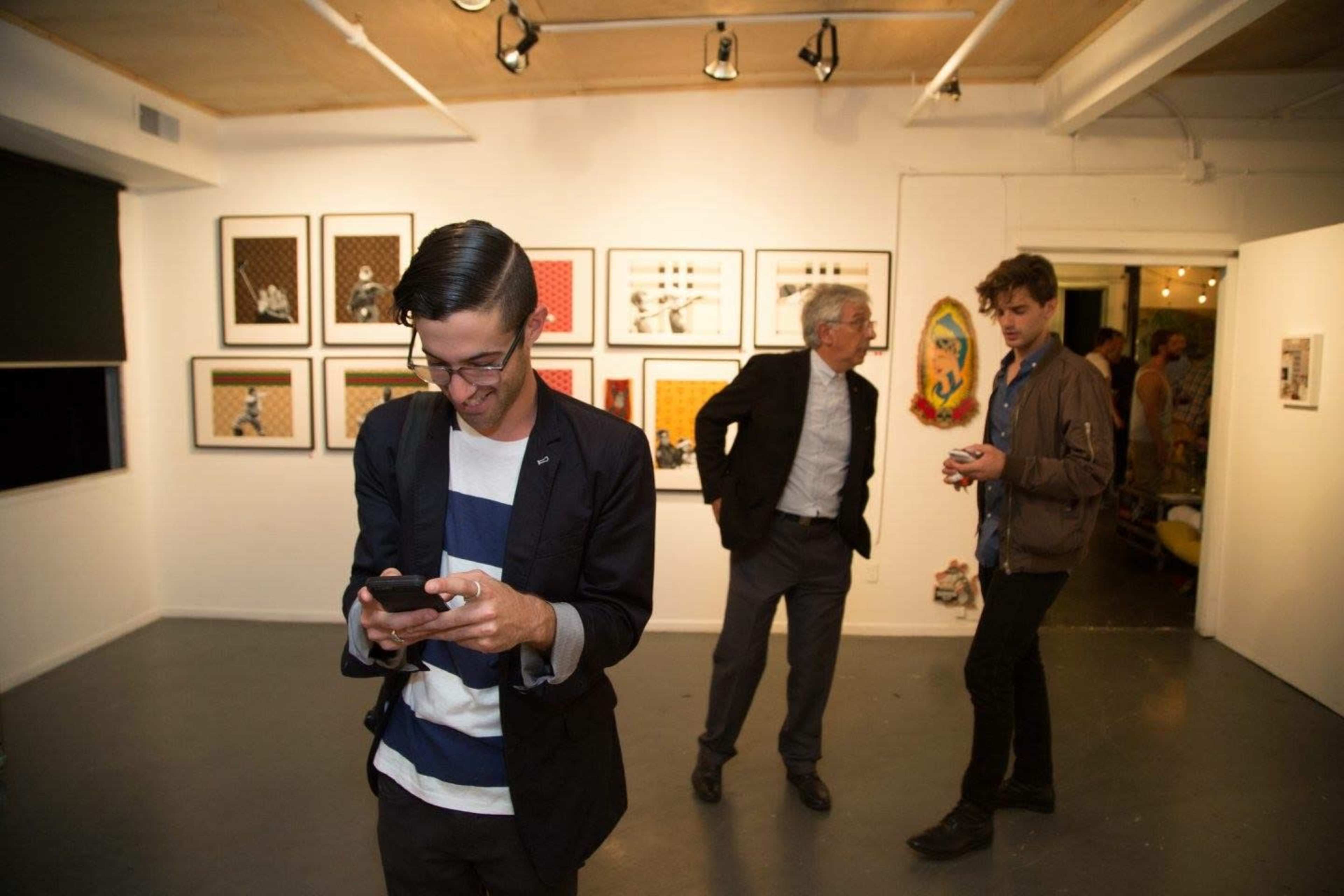 A man in a striped shirt looks at his phone while two other men converse in an art gallery surrounded by framed artwork on the walls.