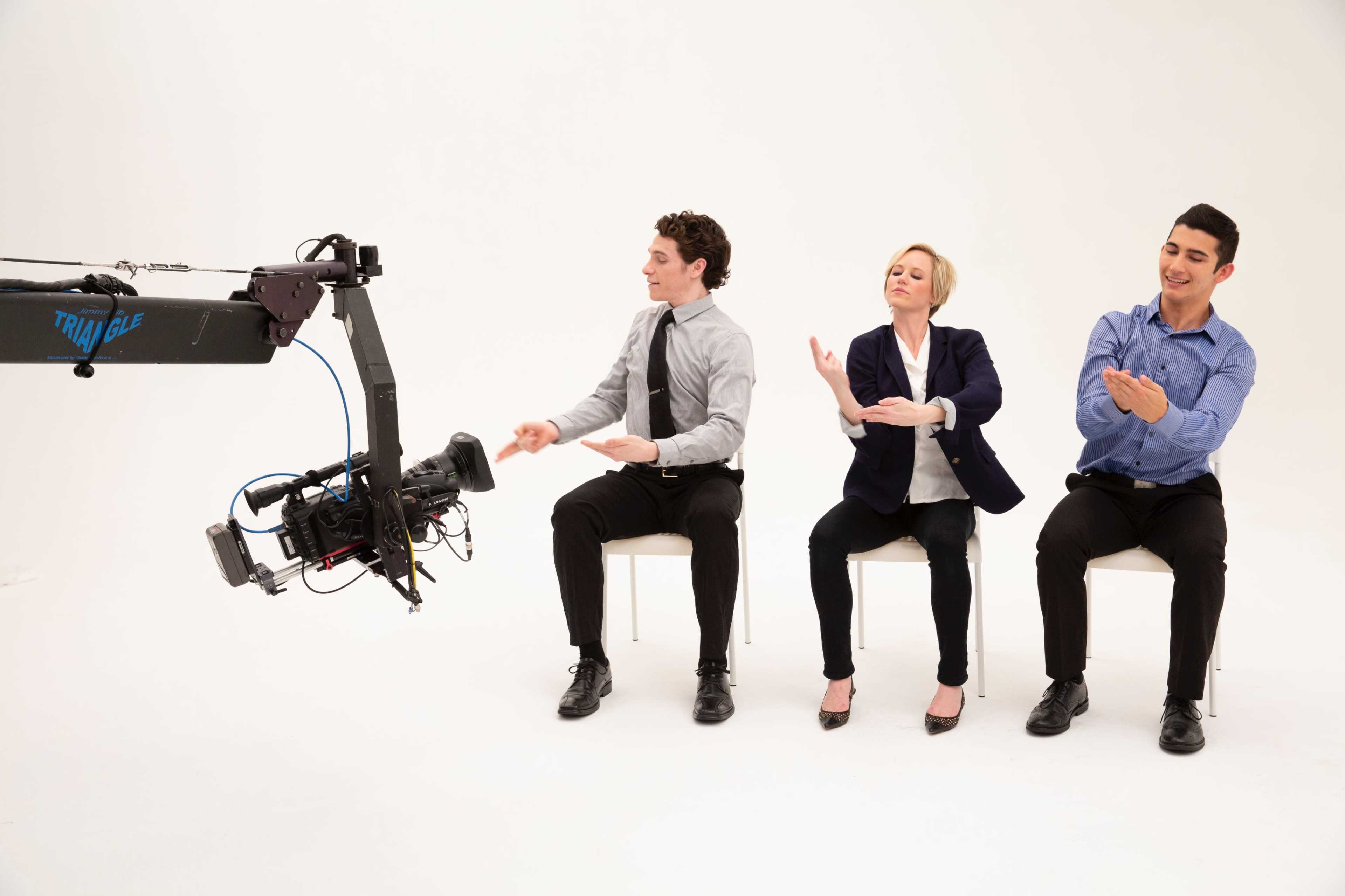 Three seated individuals face a camera on a crane, with the man on the left in a gray shirt, the woman in the center in a black blazer, and the man on the right in a blue shirt, all appearing to engage in a gestural activity.