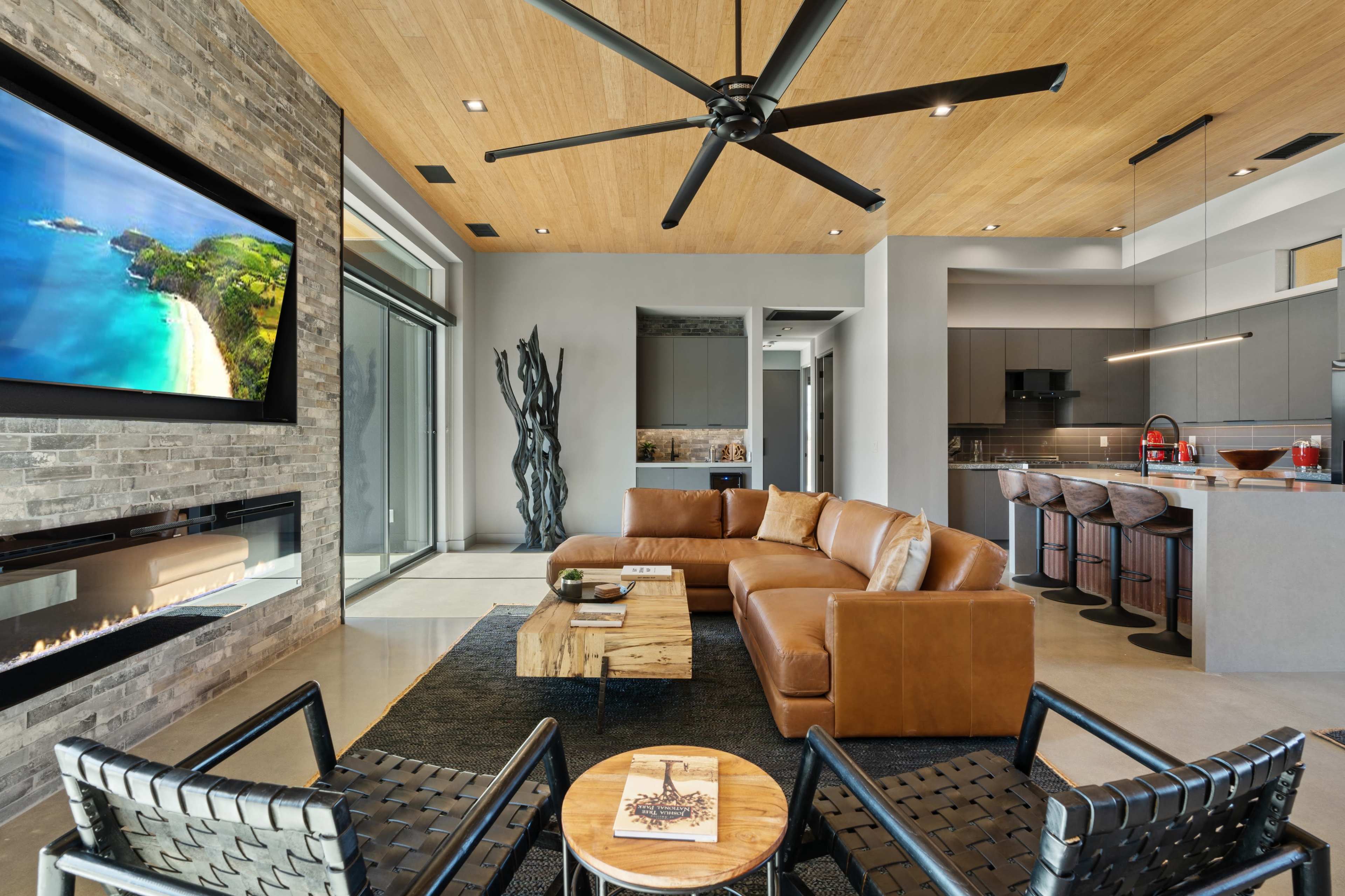 The image shows a modern living room with a large sofa, a coffee table, a flat-screen TV mounted on a stone wall, and a kitchen area in the background with bar stools.