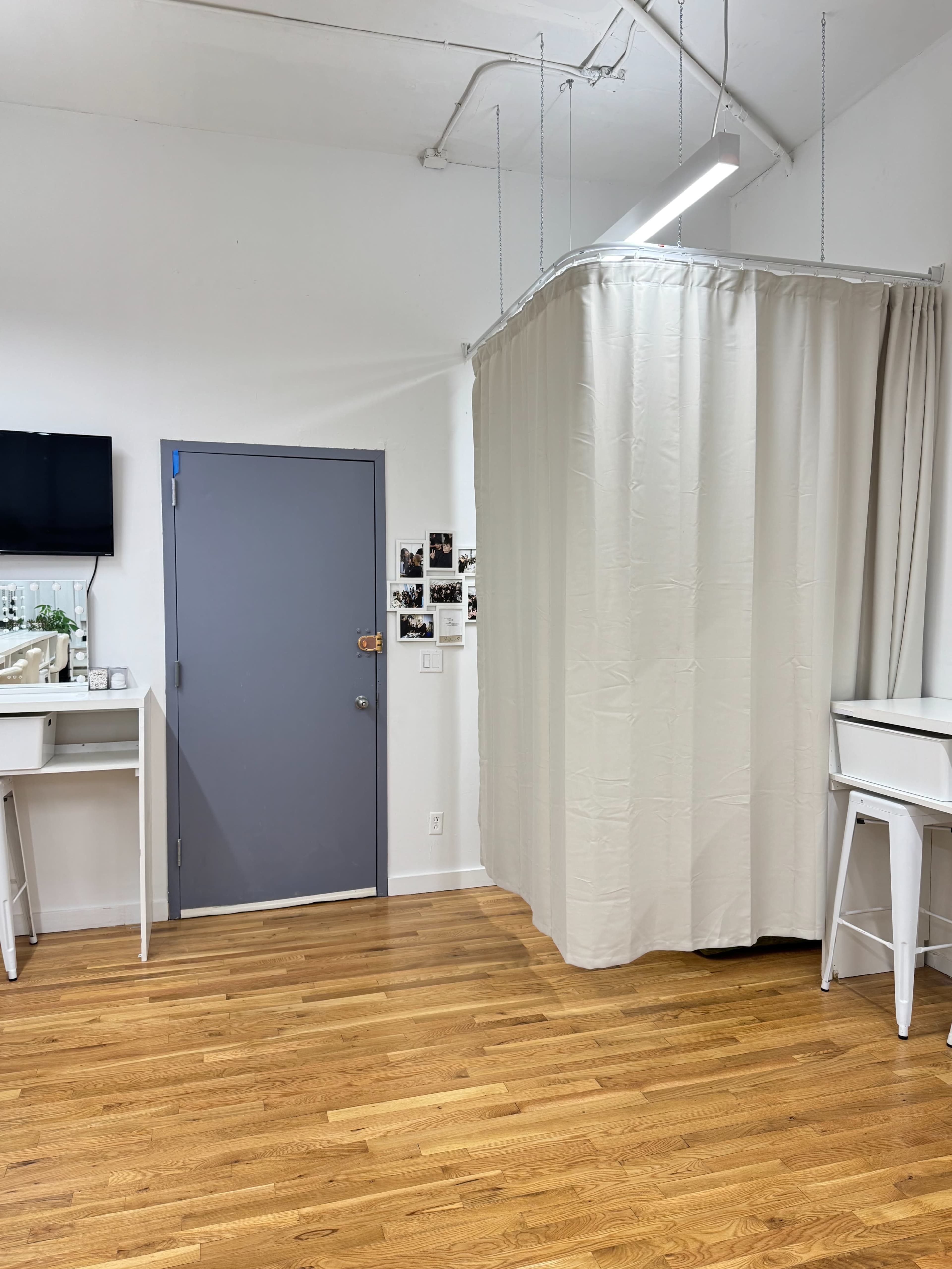 The image shows a spacious room with light wooden flooring, featuring a gray door, a wall-mounted TV, and a curtain that divides part of the space.